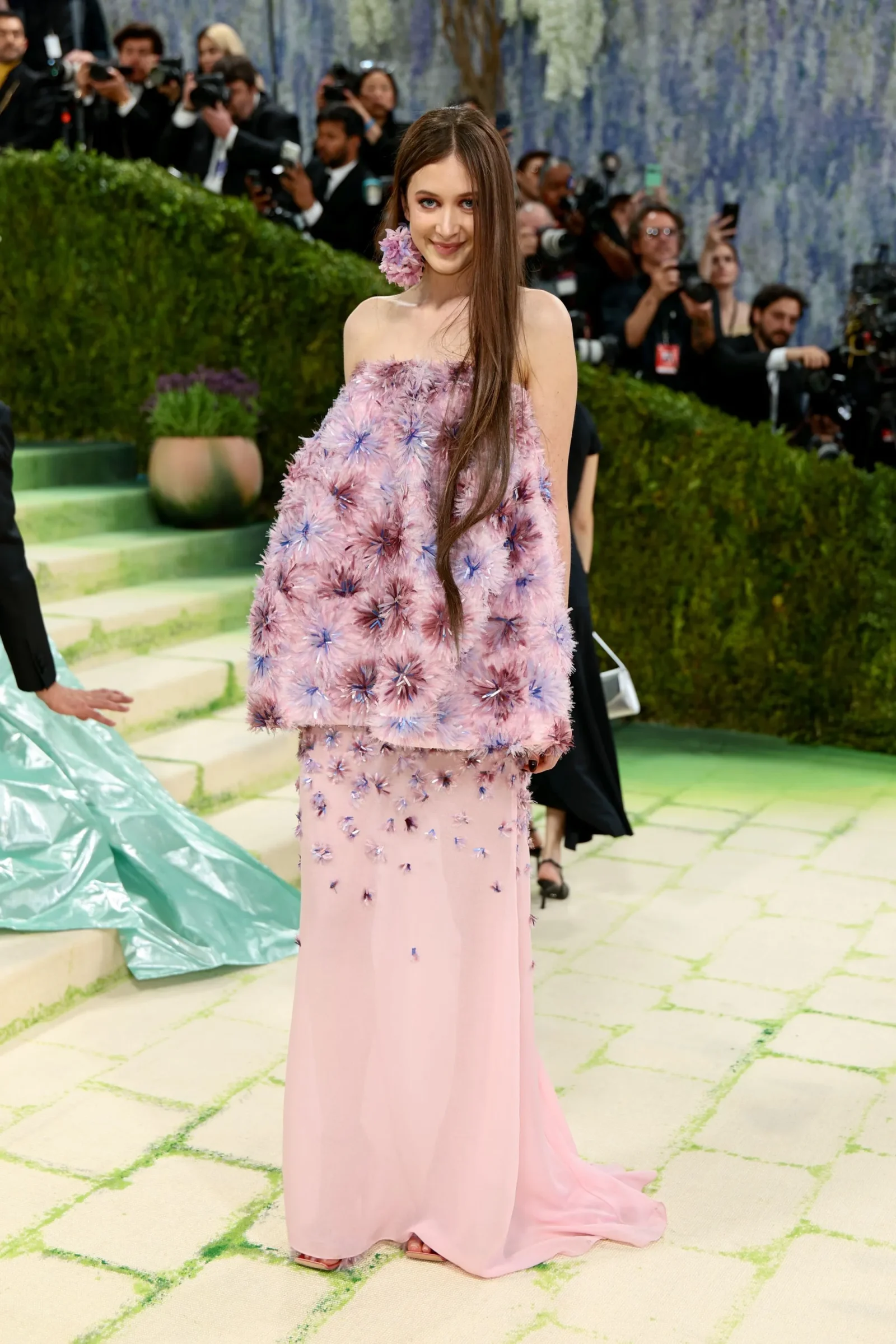 Woman in a pale pink, floral-textured gown posing on a red carpet with photographers in the background; a large flower accessory near her ear.