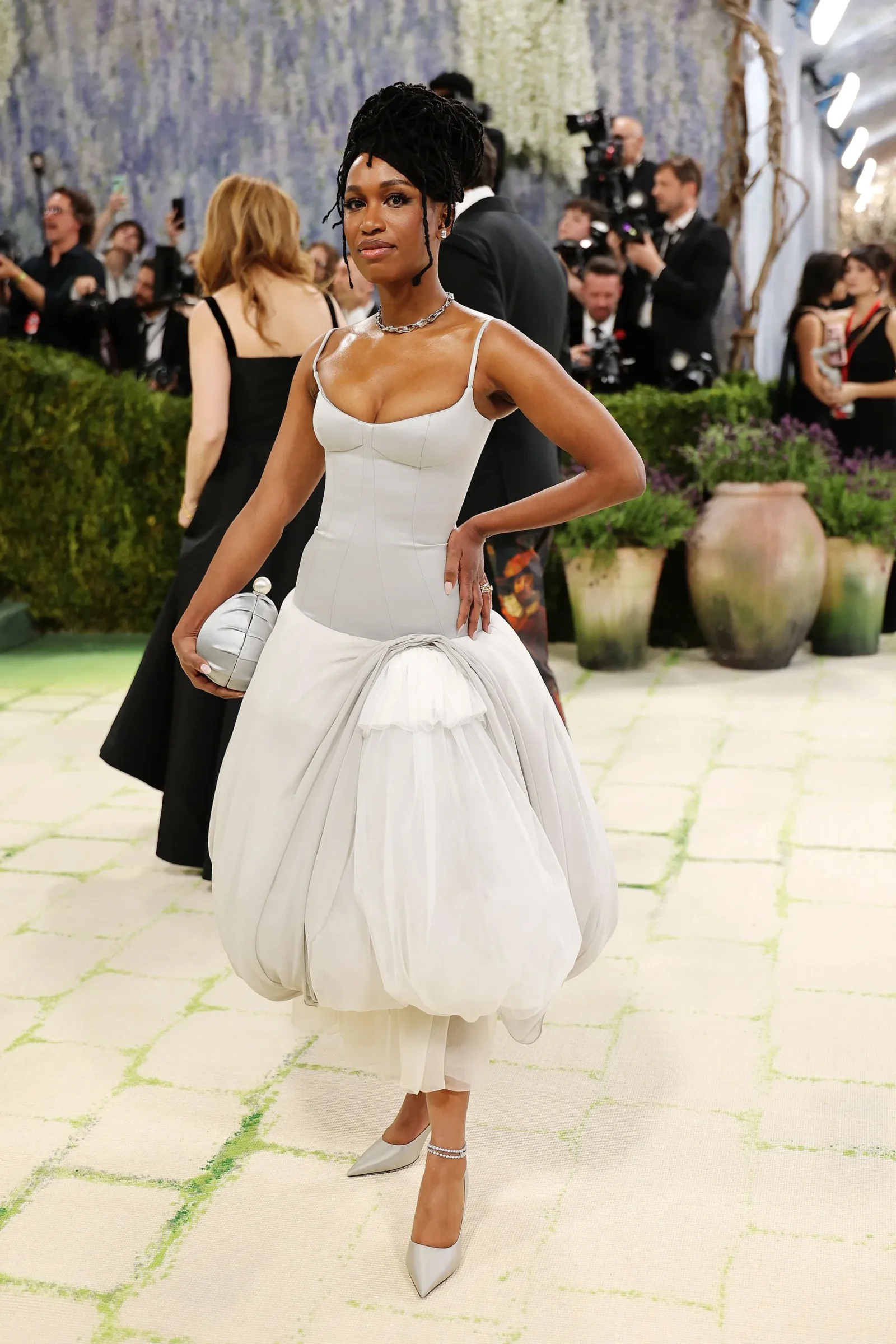 Woman in a light gray, strapless ball gown with a draped overskirt, holding a round silver clutch, on a red carpet with photographers in the background.