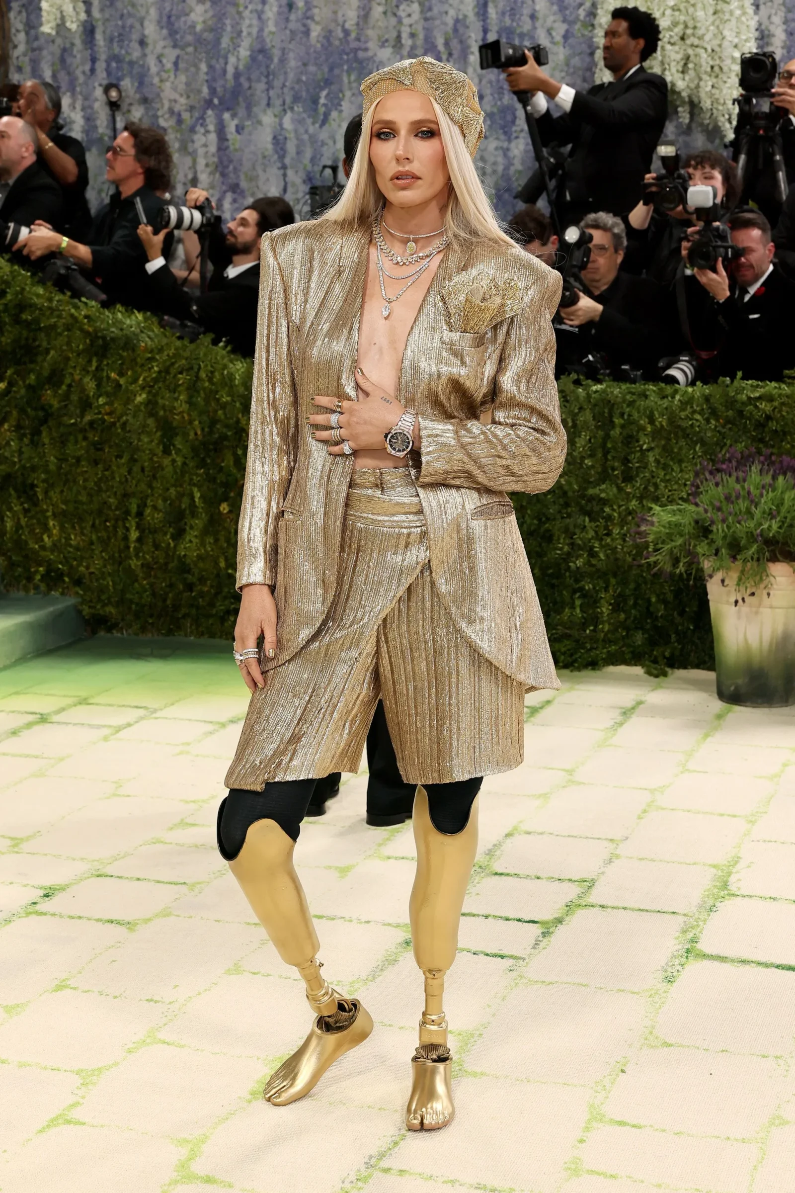 A person in a gold metallic suit and headpiece posing on a light-colored carpet with photographers in the background.