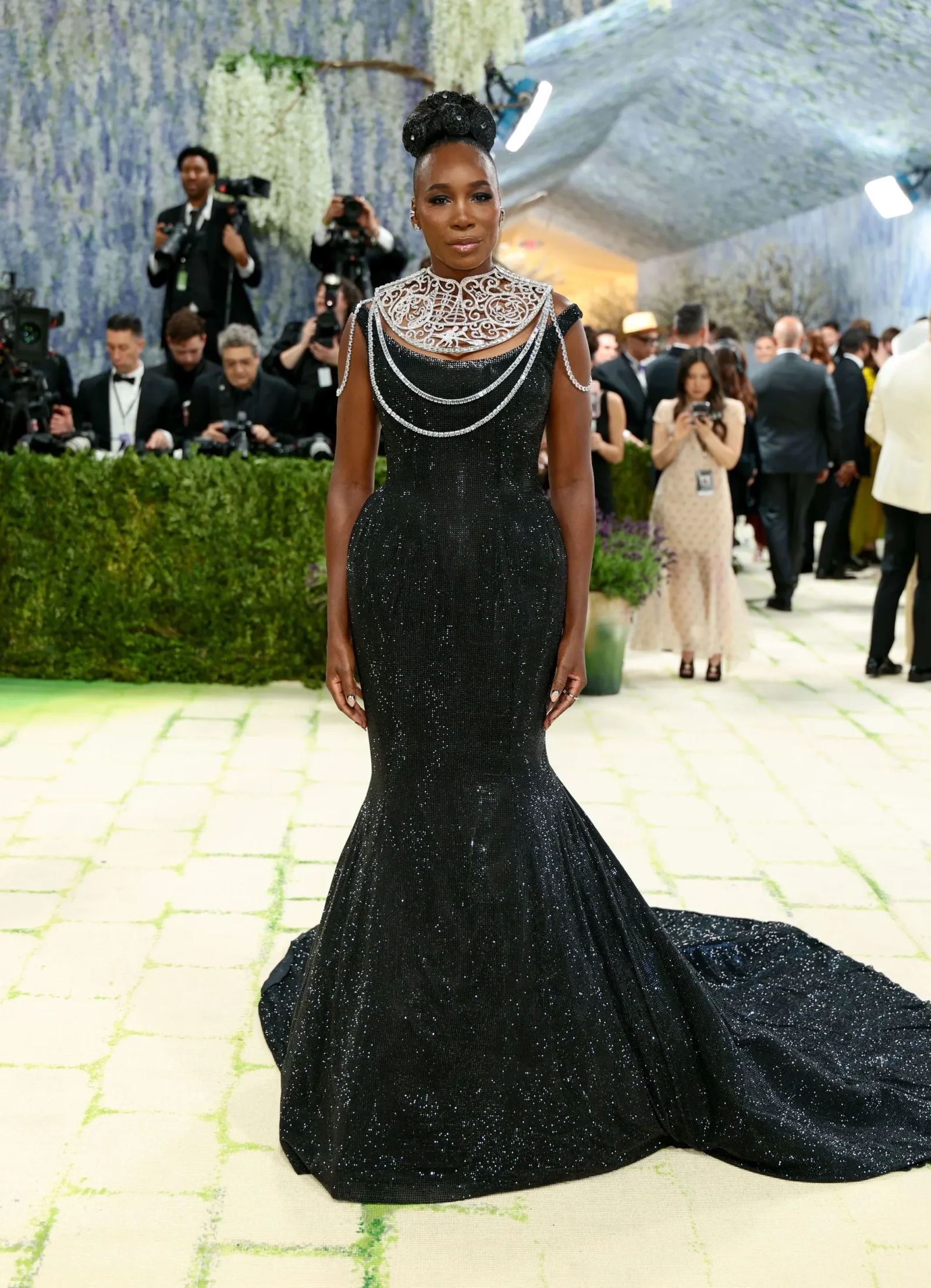 Elegant woman in a glittering black gown with a jeweled collar at a red-carpet event, photographers in the background.