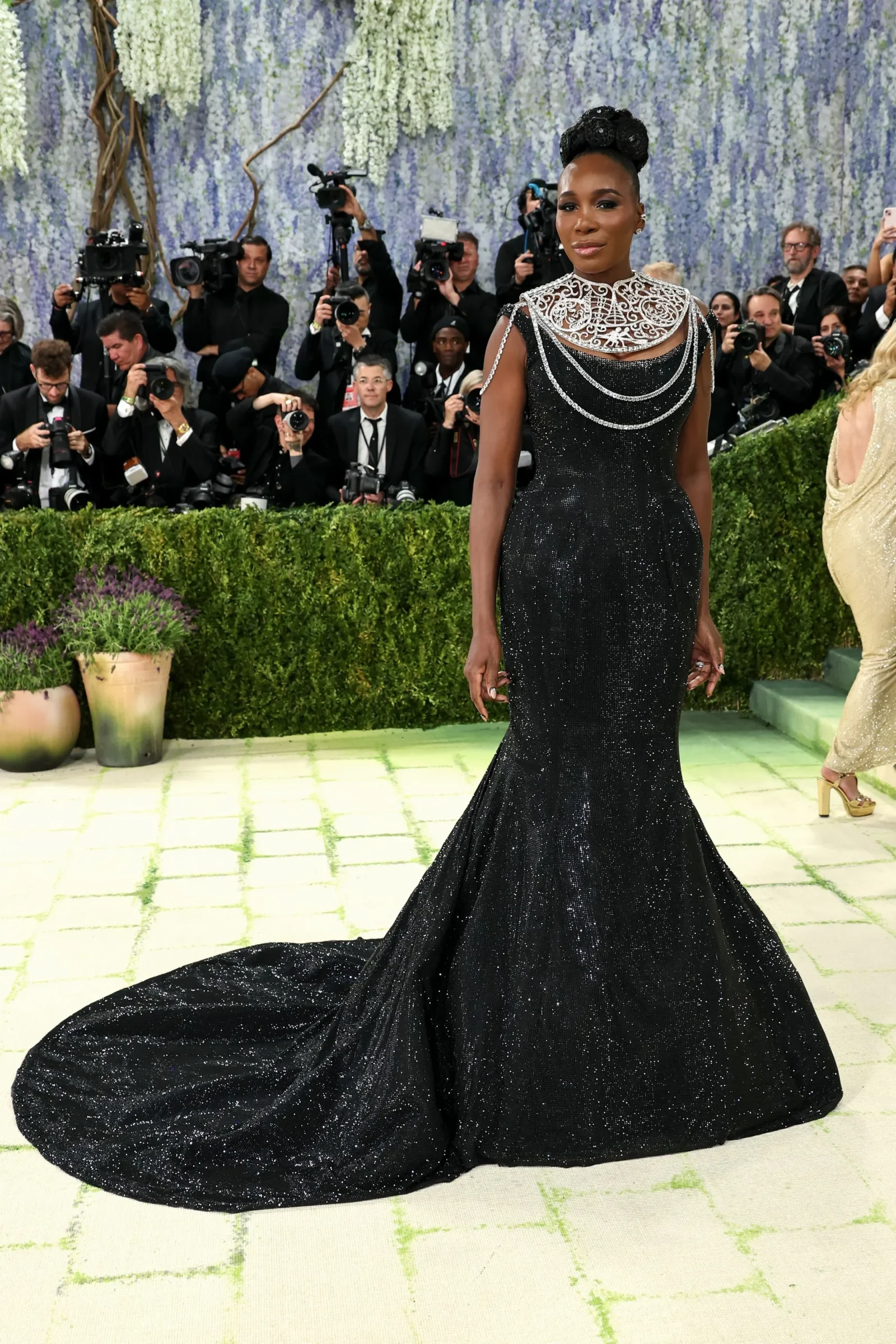 African American woman in a black glittery gown with an ornate silver necklace on a red carpet, surrounded by photographers.