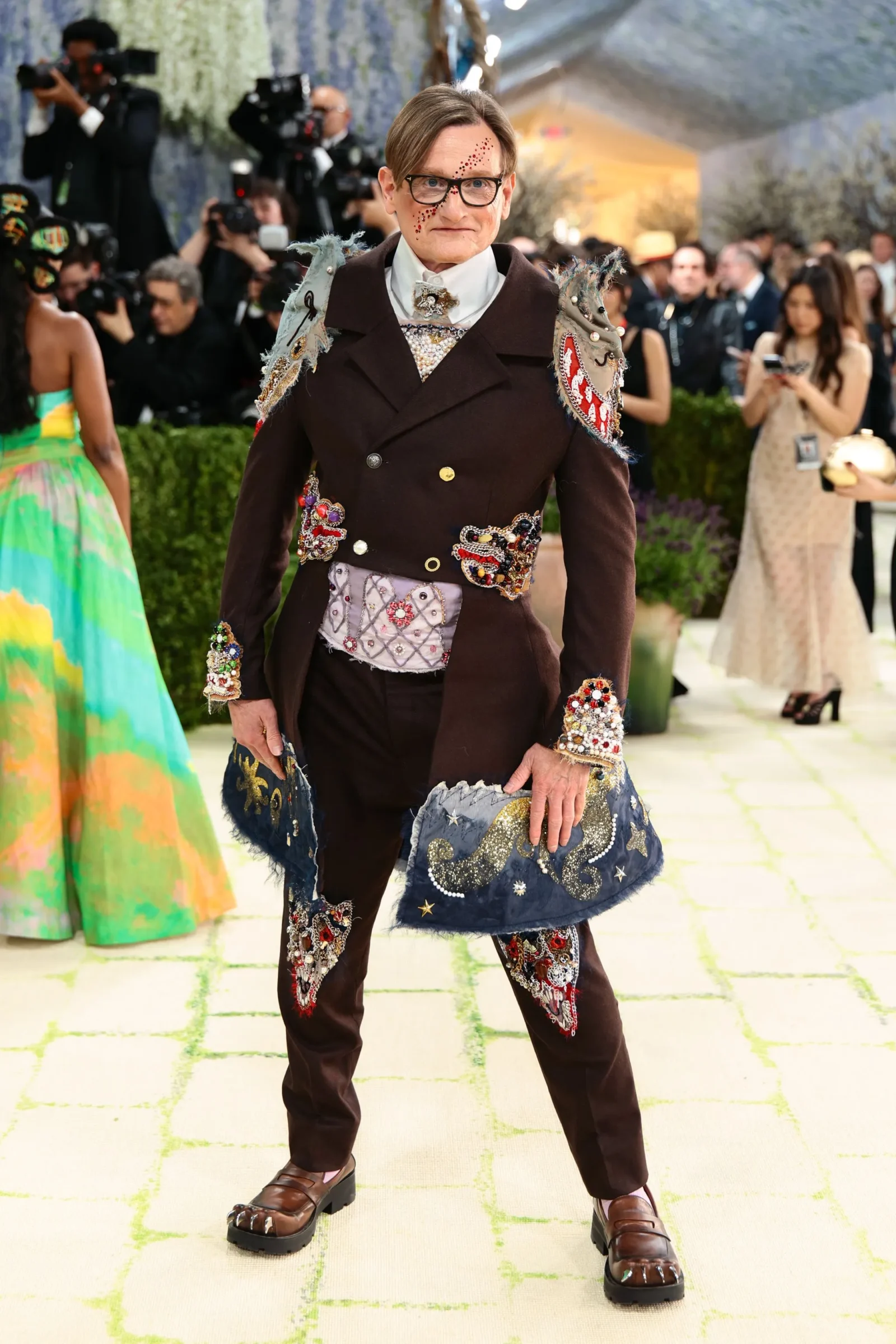 Person at a red-carpet event wearing a dark, heavily embellished coat with jeweled patches; photographers in the background.