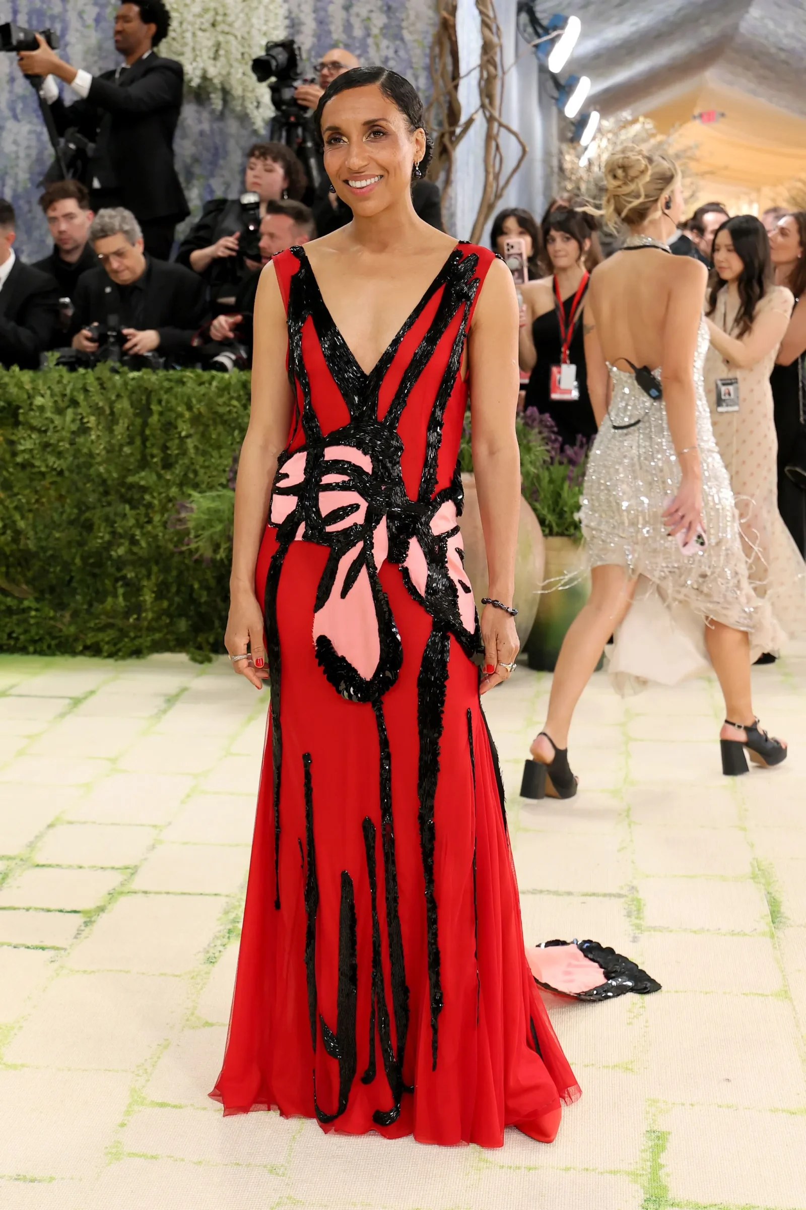 Woman wearing a red gown with black abstract patterns posing on a carpet as photographers backdrop her at a gala event.