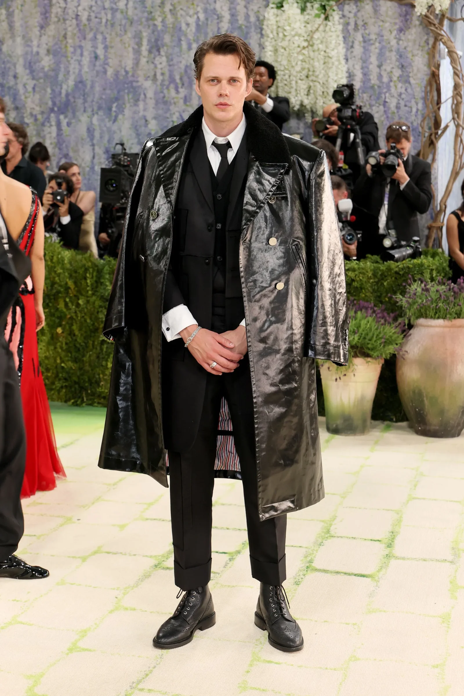Man at a red-carpet event wearing a shiny black trench coat over a black suit; photographers in the background.