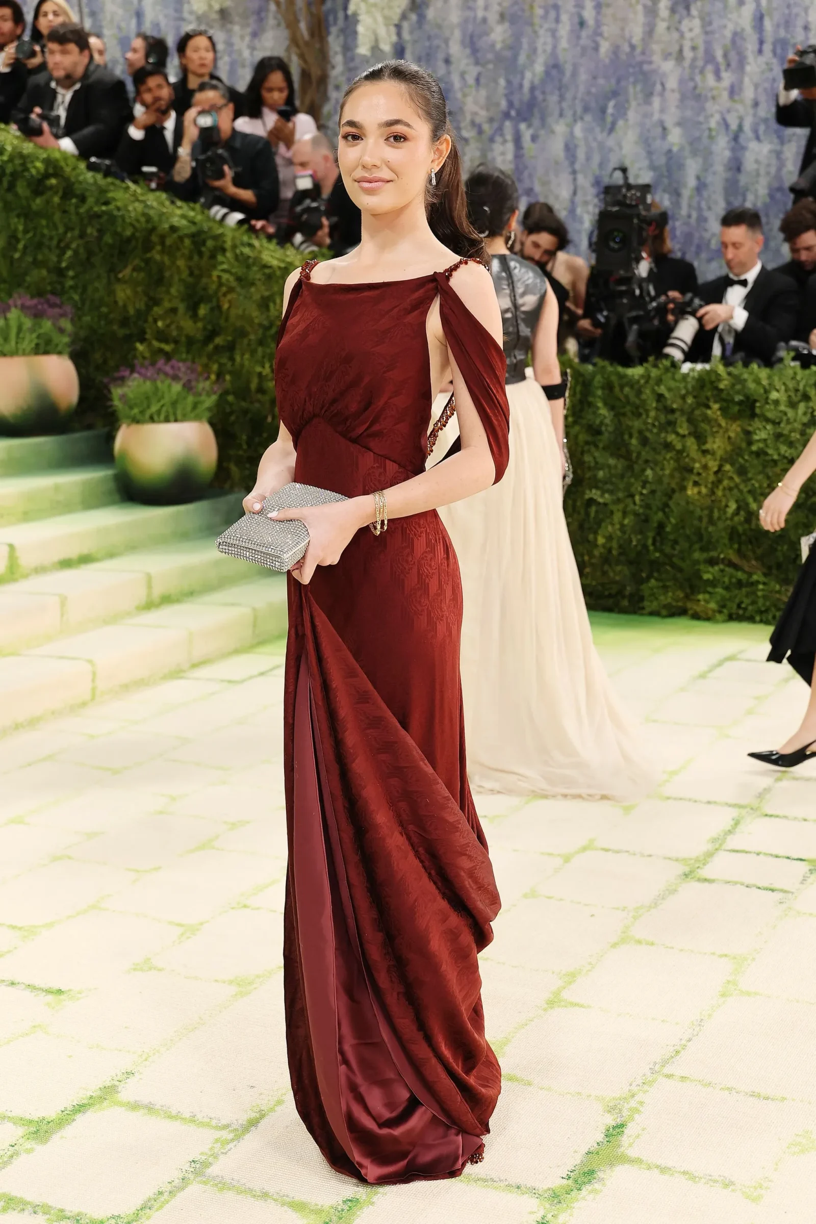 Woman in a burgundy evening gown holding a silver clutch on a red carpet, with photographers in the background.