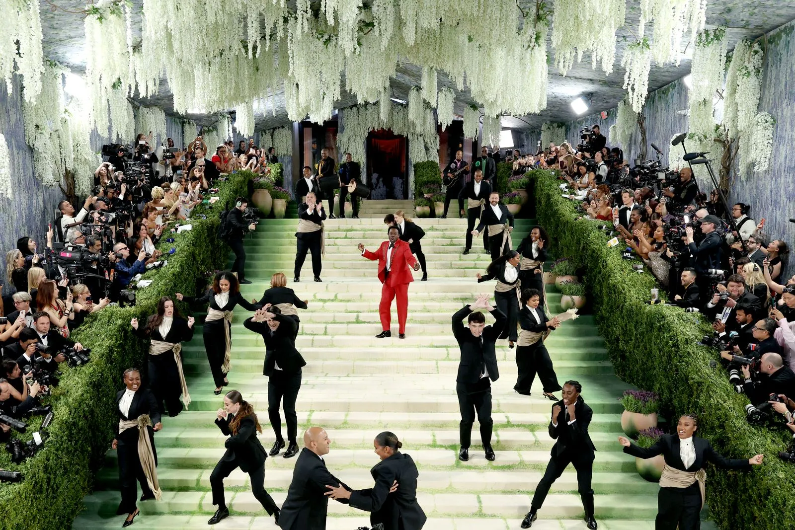 A performer in a bright red suit dances on a pale-green staircase surrounded by dancers in black outfits with beige sashes as paparazzi line both sides.