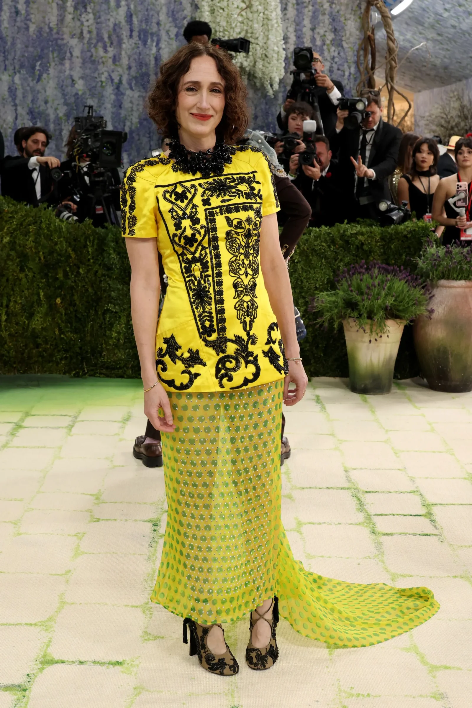 Woman posing on a lime-green carpet in a bright yellow embroidered top and green patterned skirt, photographers and cameras in the background.