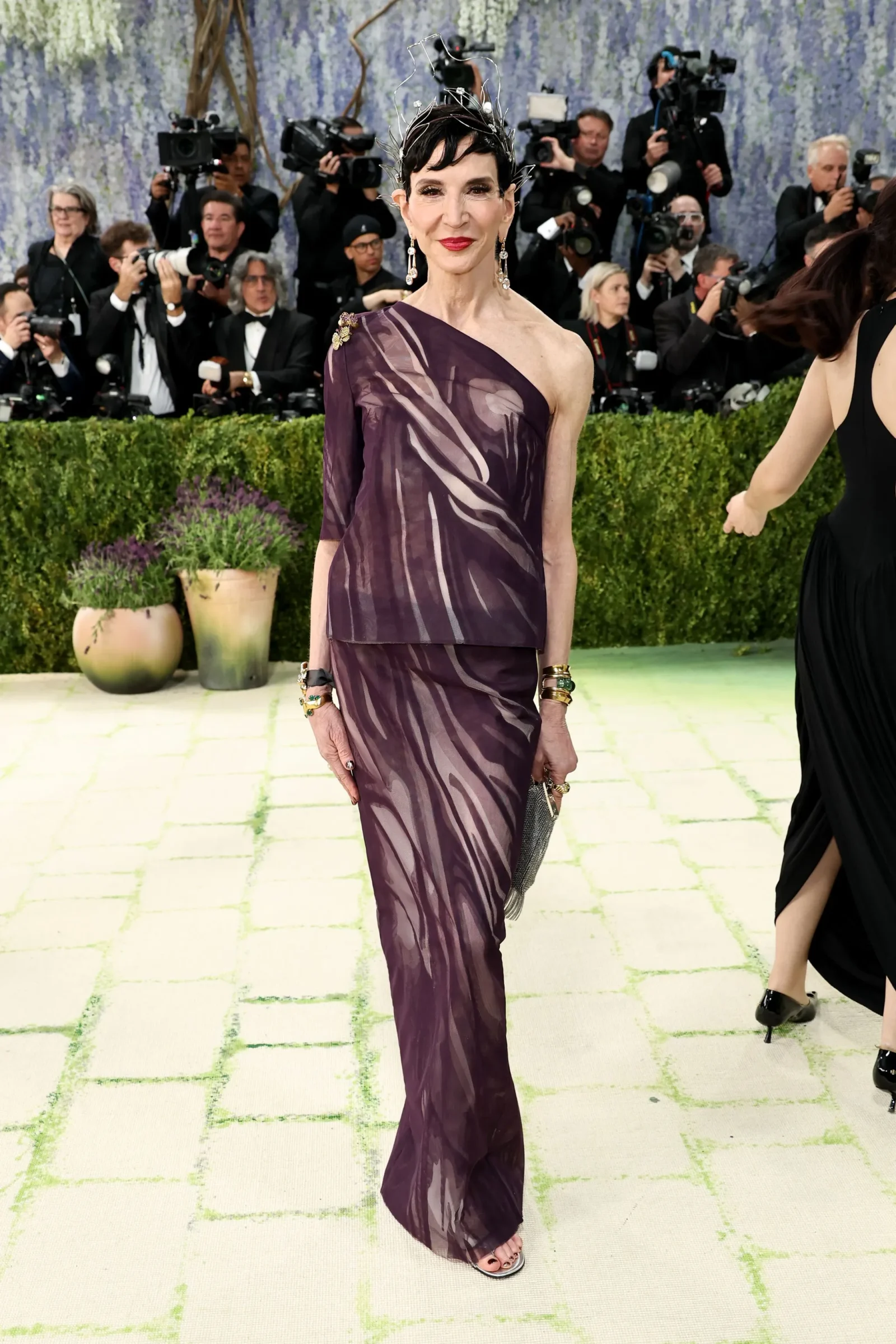 Elegant woman in a purple, one-shoulder gown posing on a light-green carpet at a formal event, with numerous photographers in the background.