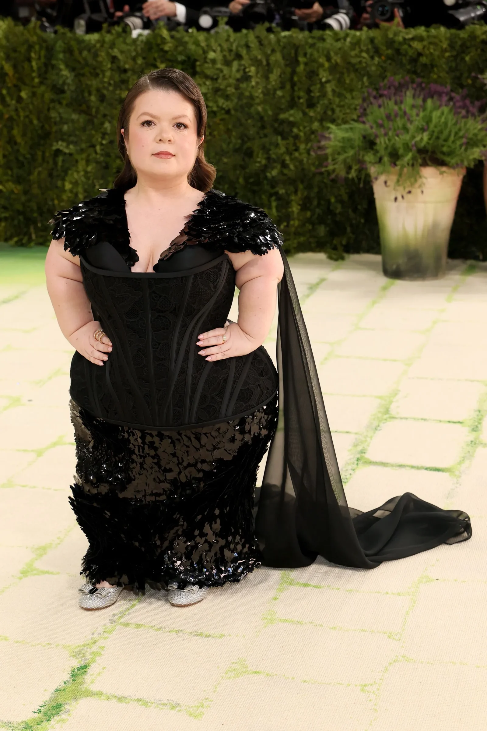Woman in a black sequined gown posing on a light carpet with photographers in the background at a formal event.