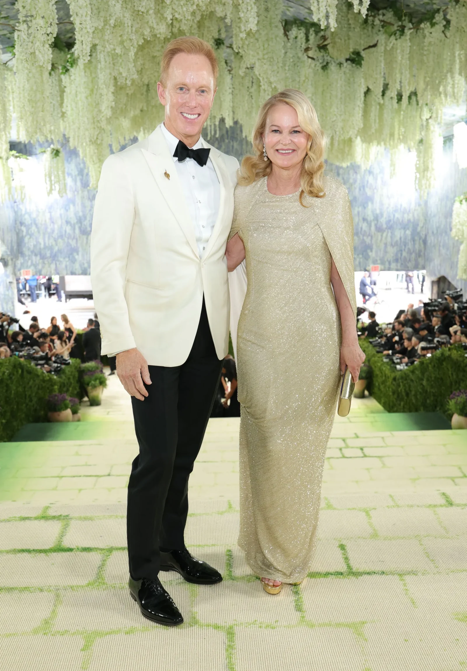 Smiling man in a white tuxedo with black bow tie stands next to a smiling woman in a gold sequined gown under hanging white flowers.