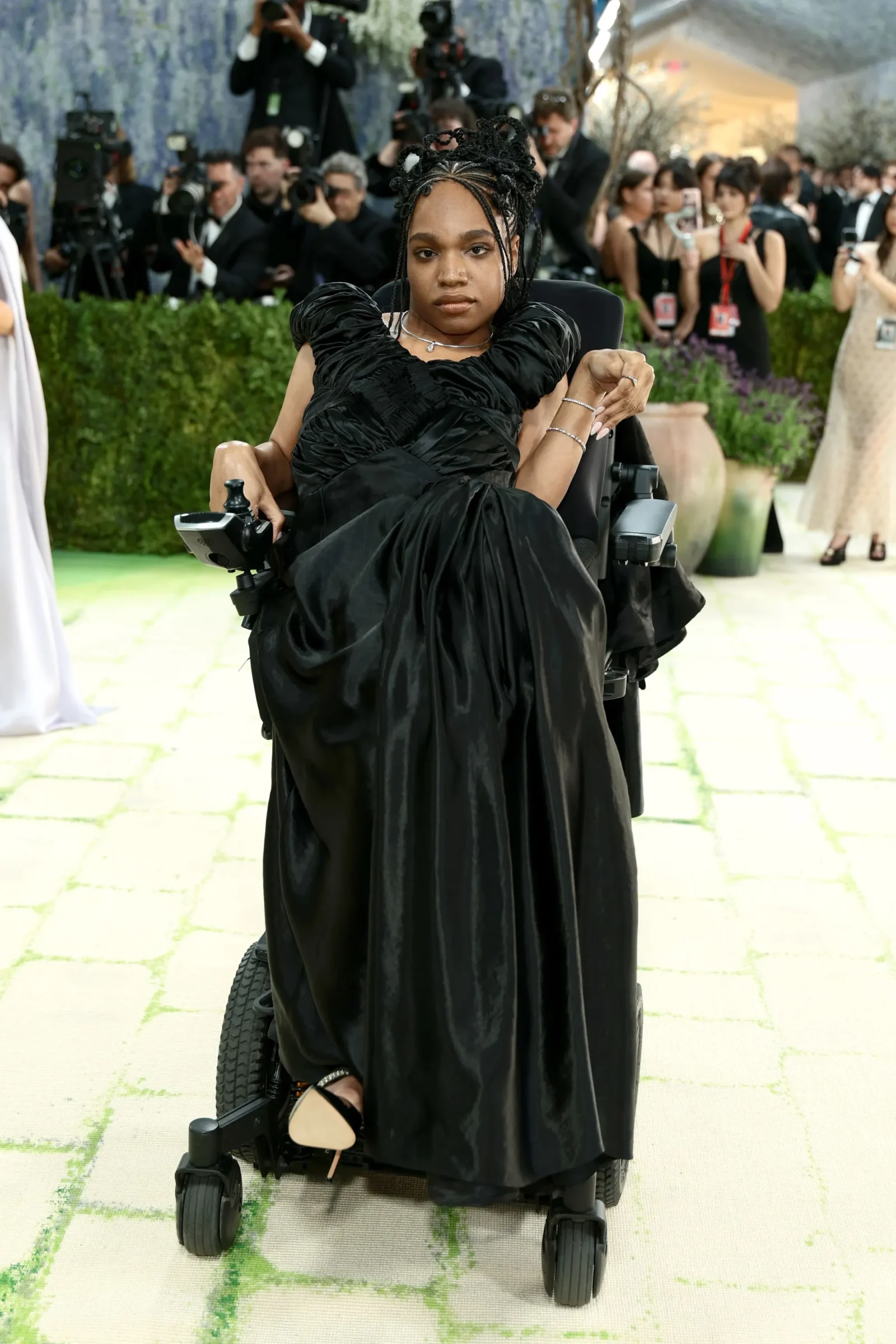 Woman in a black formal gown seated in a motorized wheelchair at a red-carpet-style event, photographers in the background.