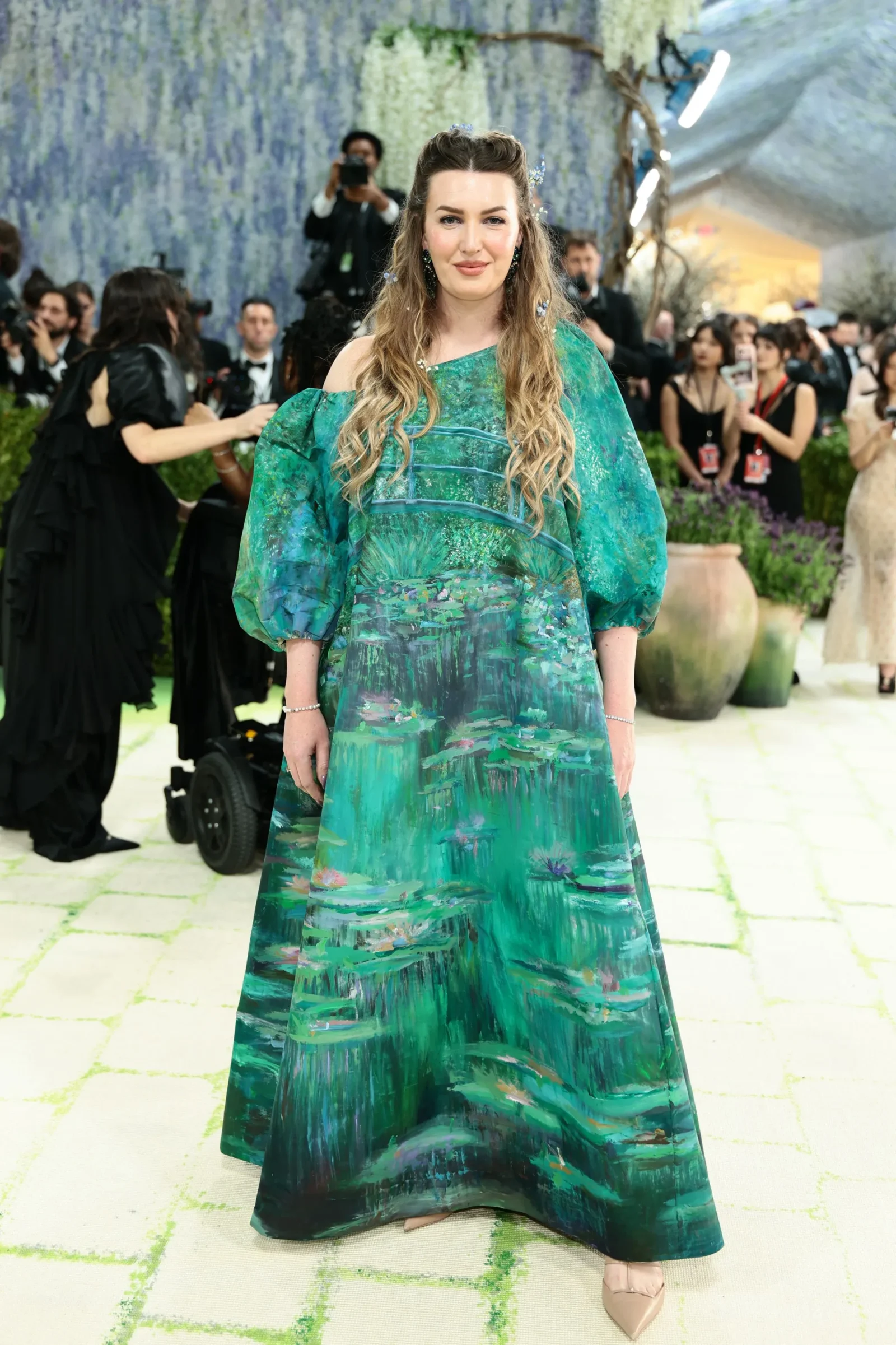 Woman in a teal-green patterned off-shoulder gown standing on a light-colored runway with a floral blue backdrop and photographers in the background