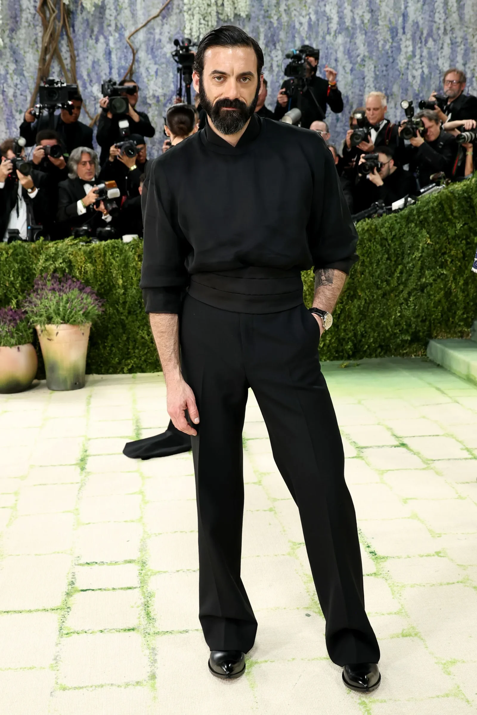 Bearded man in black outfit posing on a tiled floor as paparazzi photograph him in the background at a red carpet event.