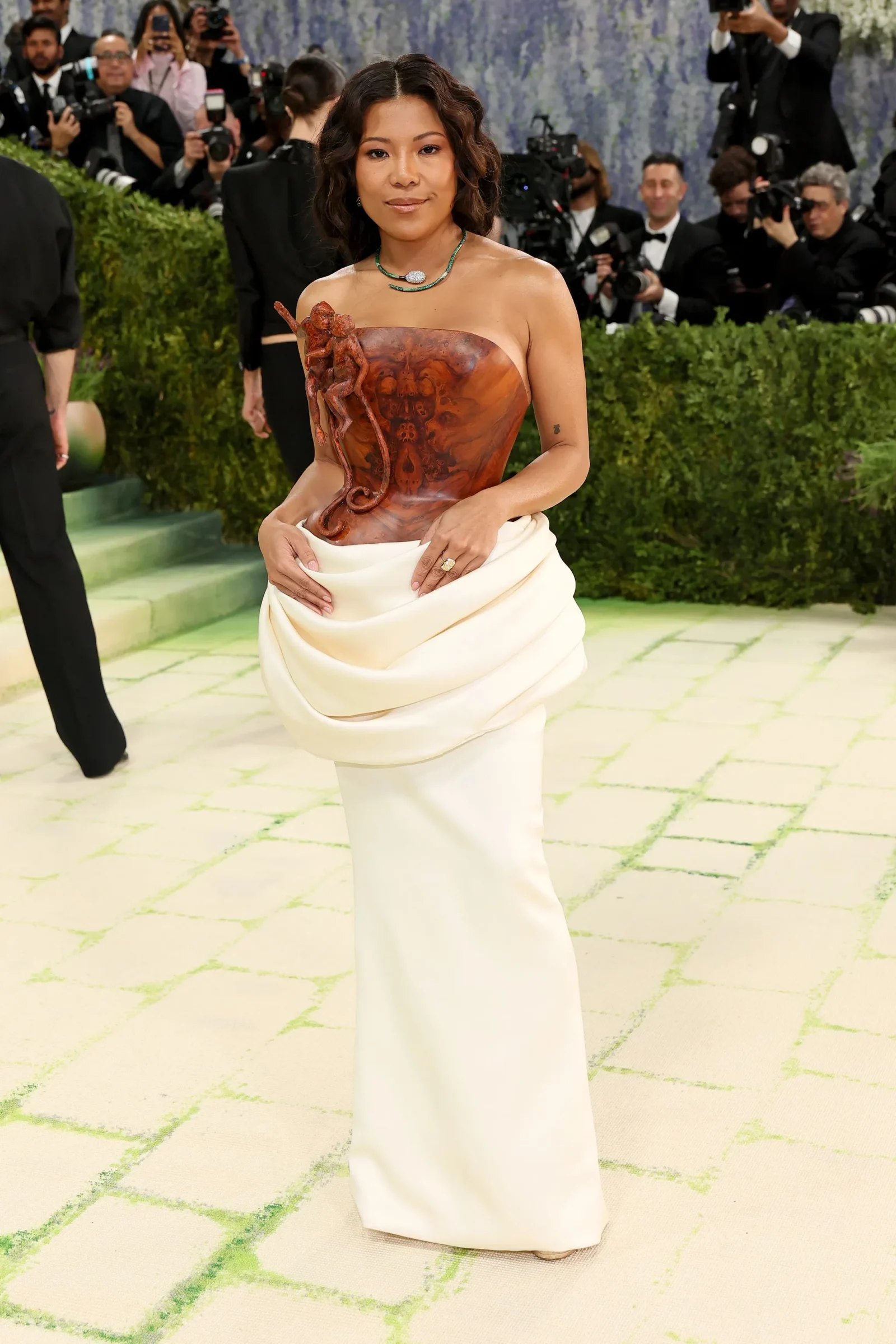 Woman on a red carpet wearing a strapless rust-toned sculptural bodice and a draped white skirt, photographers in the background.