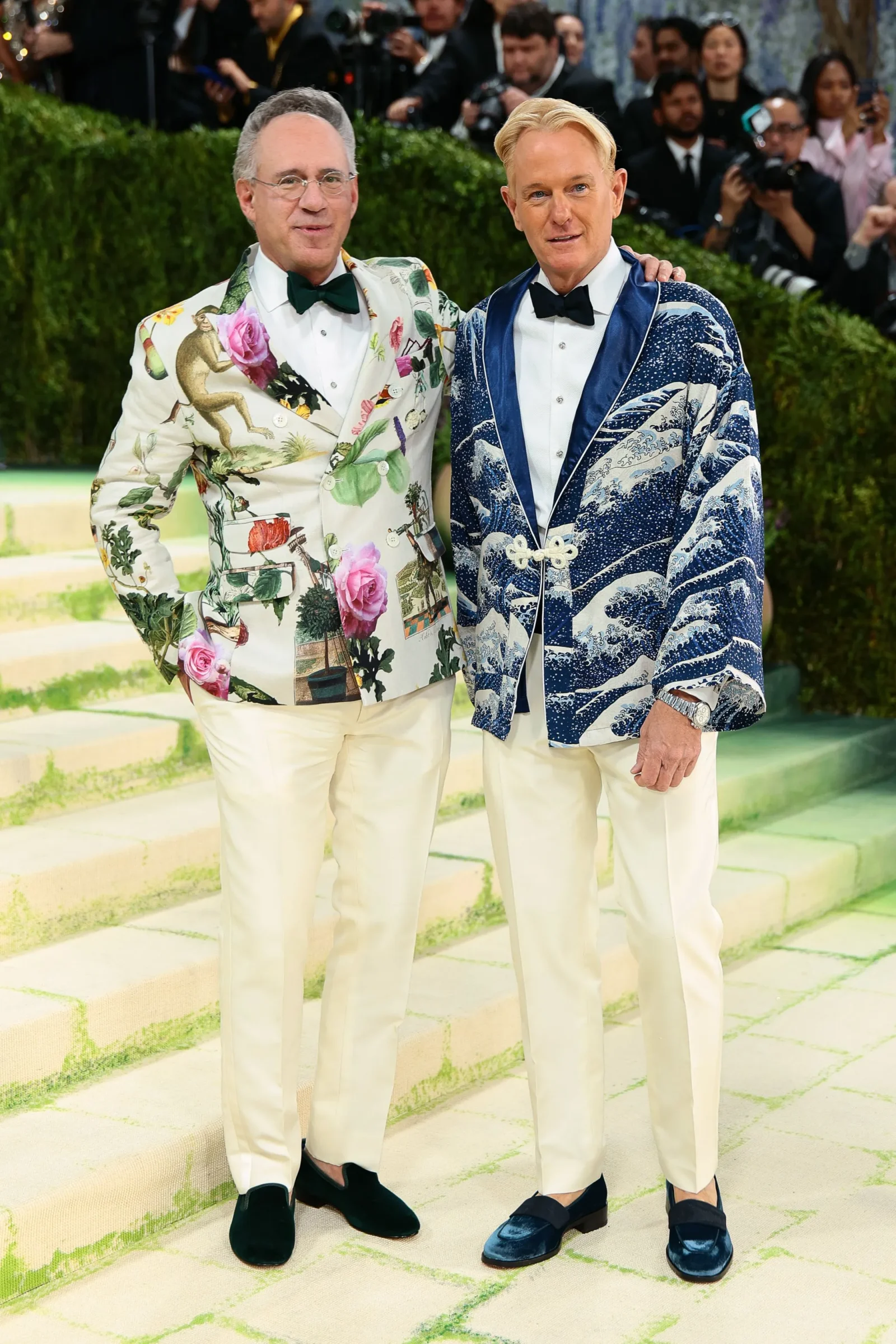 Two men pose together on stone steps at a gala, wearing bold patterned tuxedos and bow ties as photographers look on in the background.