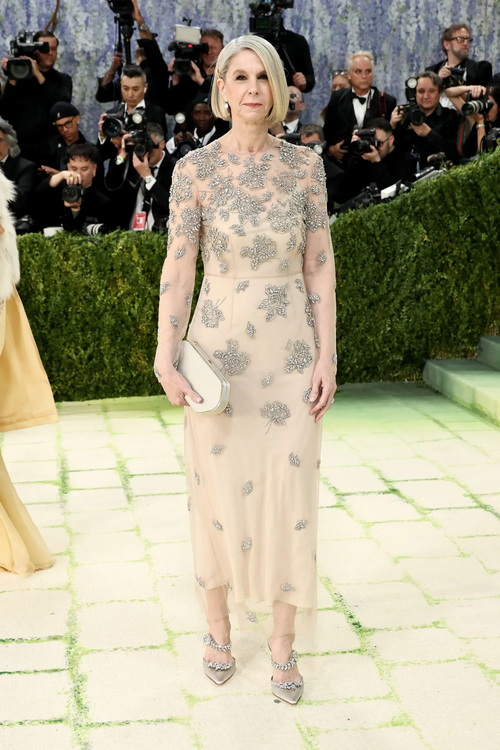 Woman in a pale pink gown with silver floral embroidery posing on a red carpet as photographers surround her in the background, holding a light clutch.