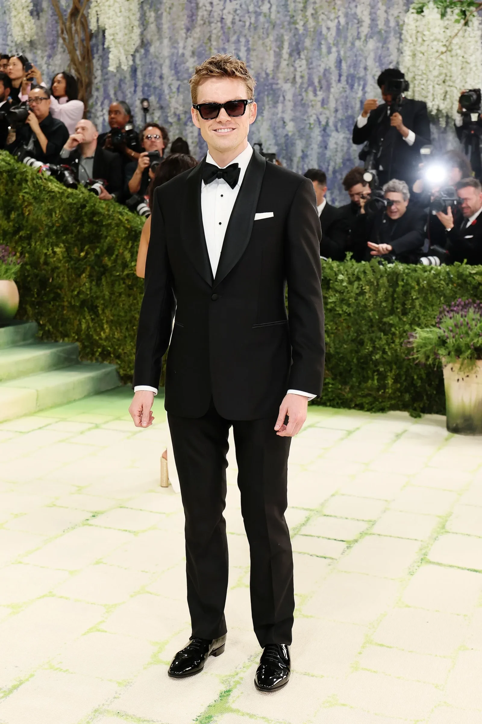 Man in a black tuxedo with bow tie posing on a light-colored carpet as photographers crowd the background.
