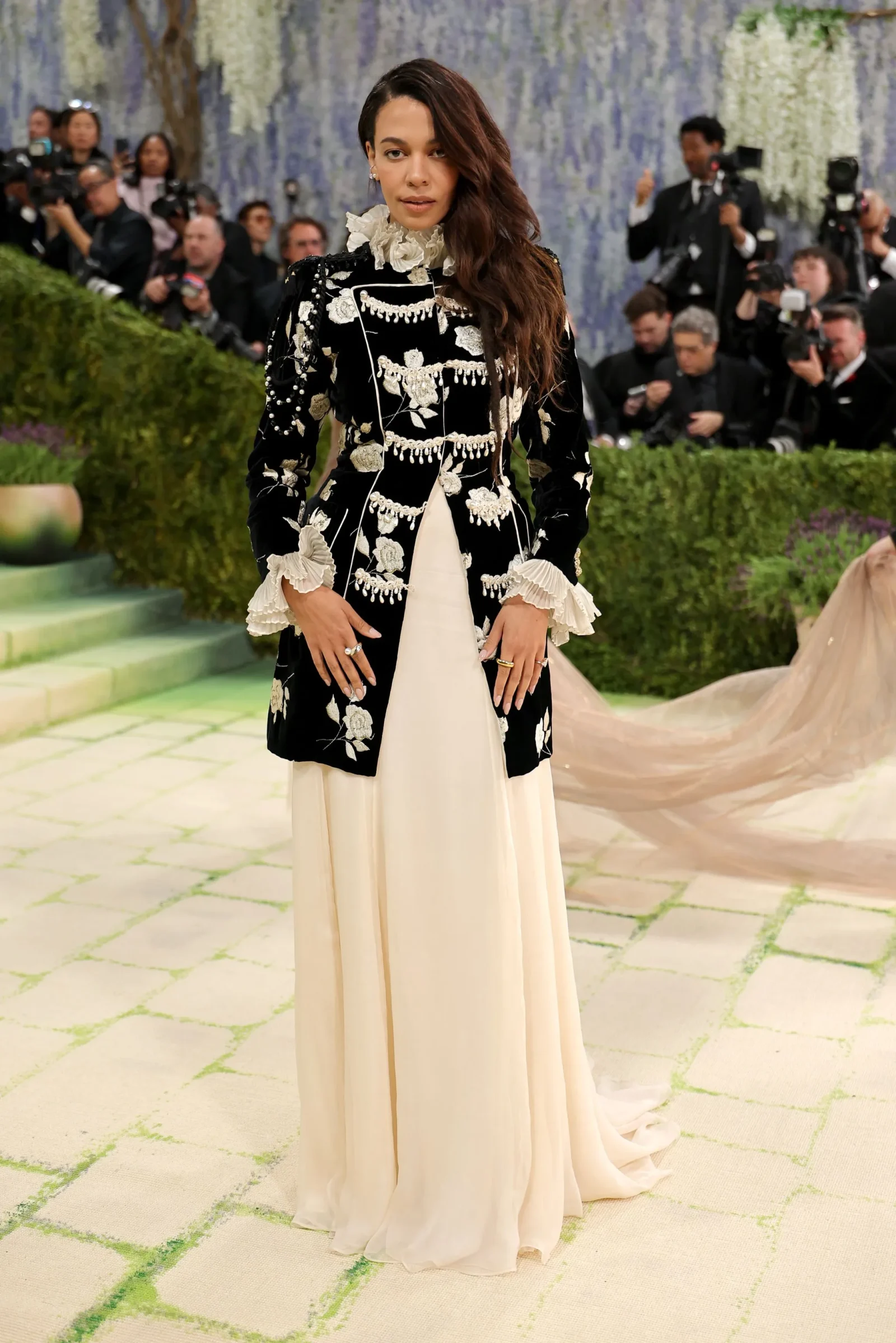 Woman in a black ornate embroidered jacket over a cream gown at a formal event, photographers in the background.