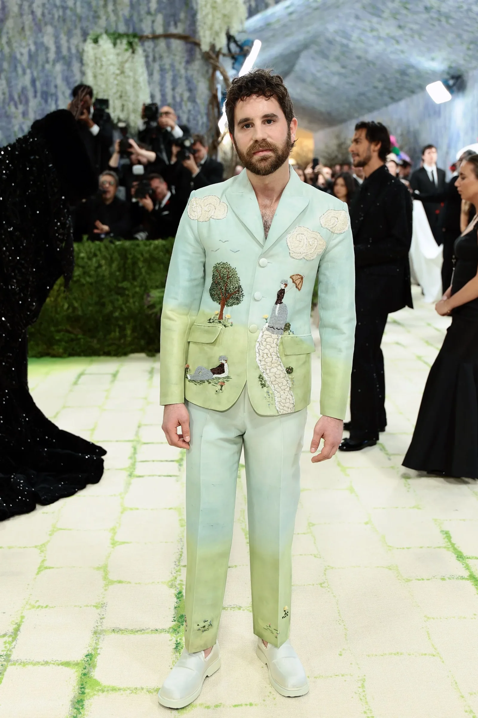 Man on a red carpet wearing a pale green suit with embroidered trees, clouds, and a winding path; photographers in the background.