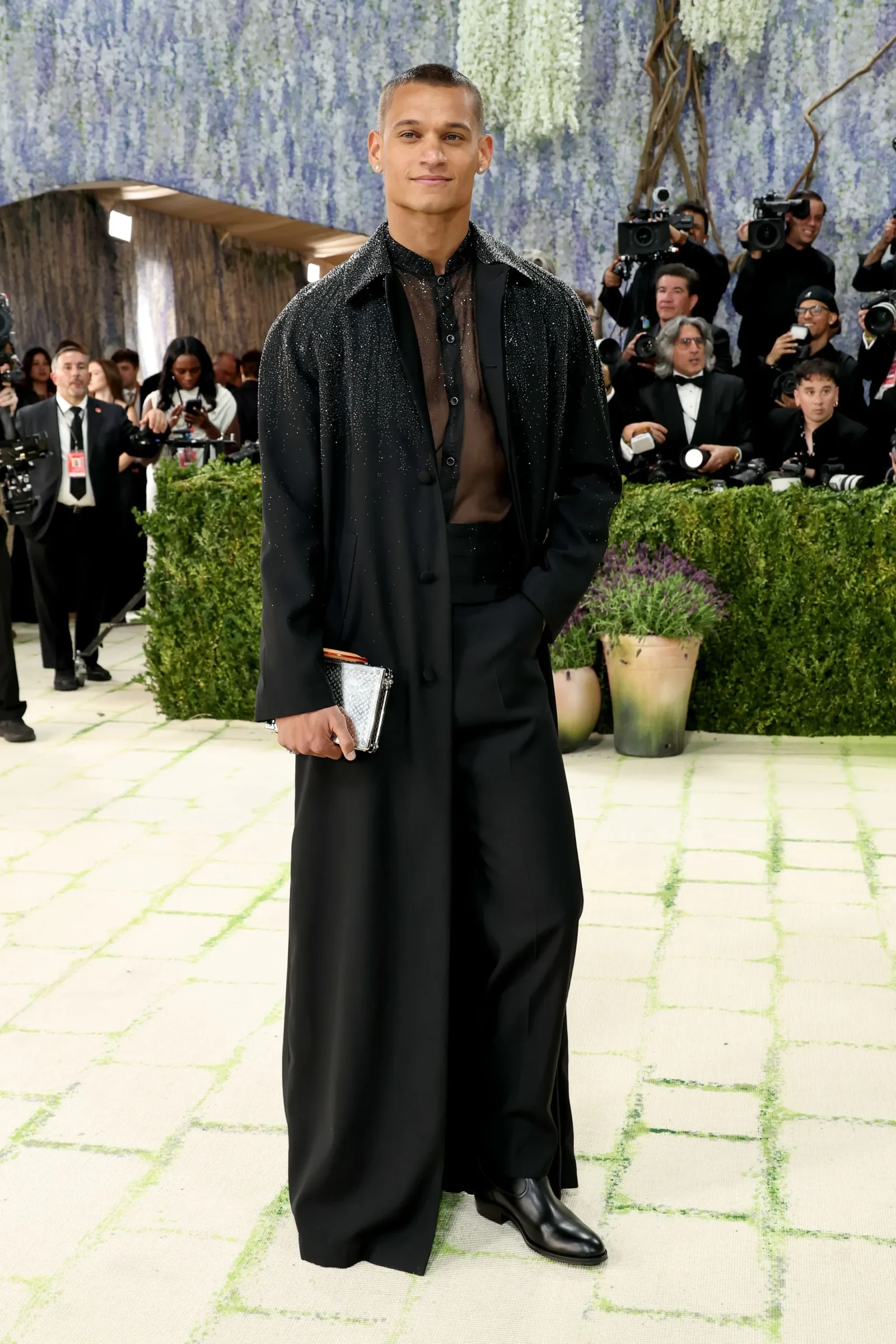 Male model on a red carpet wearing a black sheer top and long black coat, holding a silver clutch, with photographers and a floral backdrop behind.