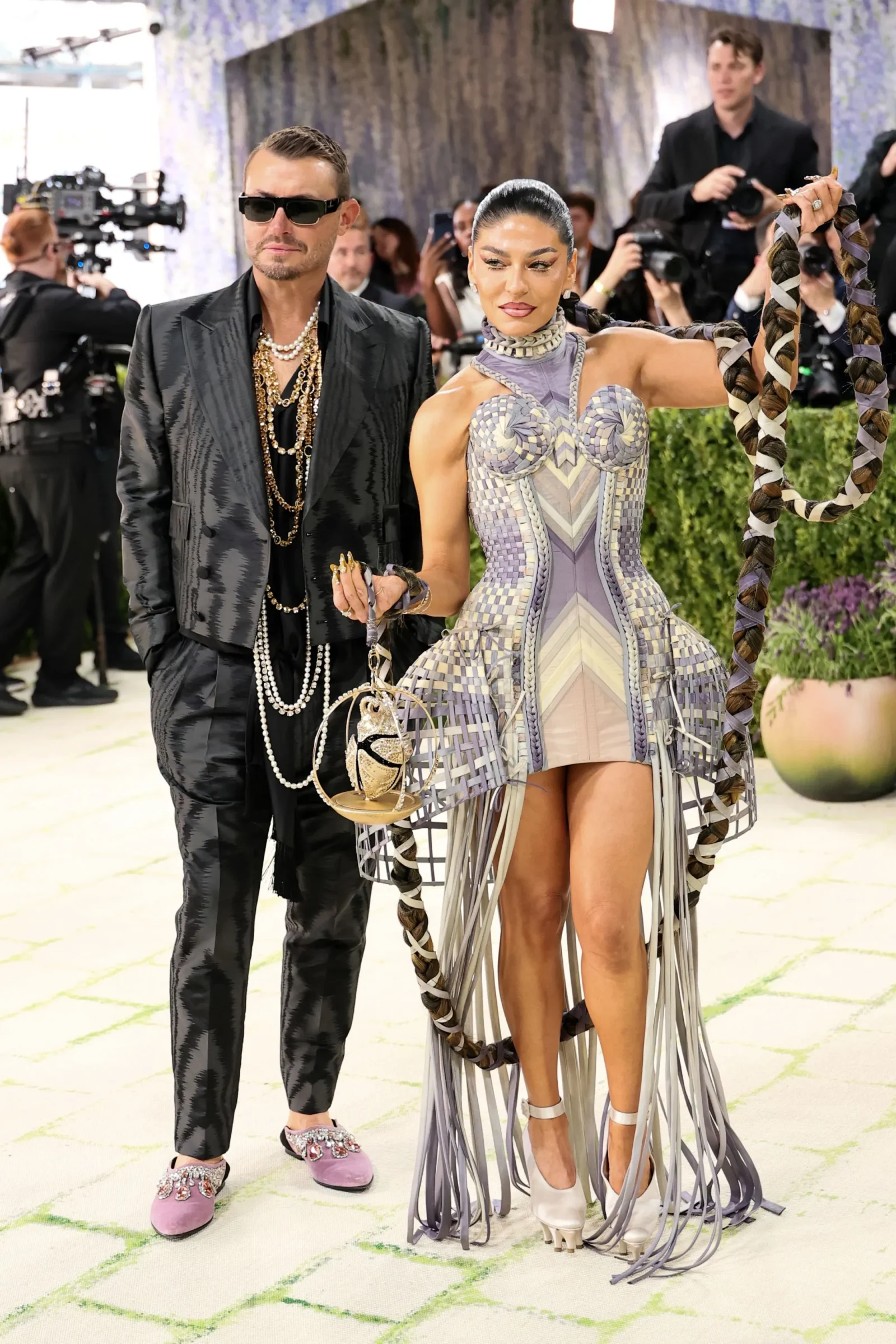 Man in a black patterned suit with pearl chains and sunglasses beside a woman in a lavender geometric dress with long fringe, at a red carpet event.