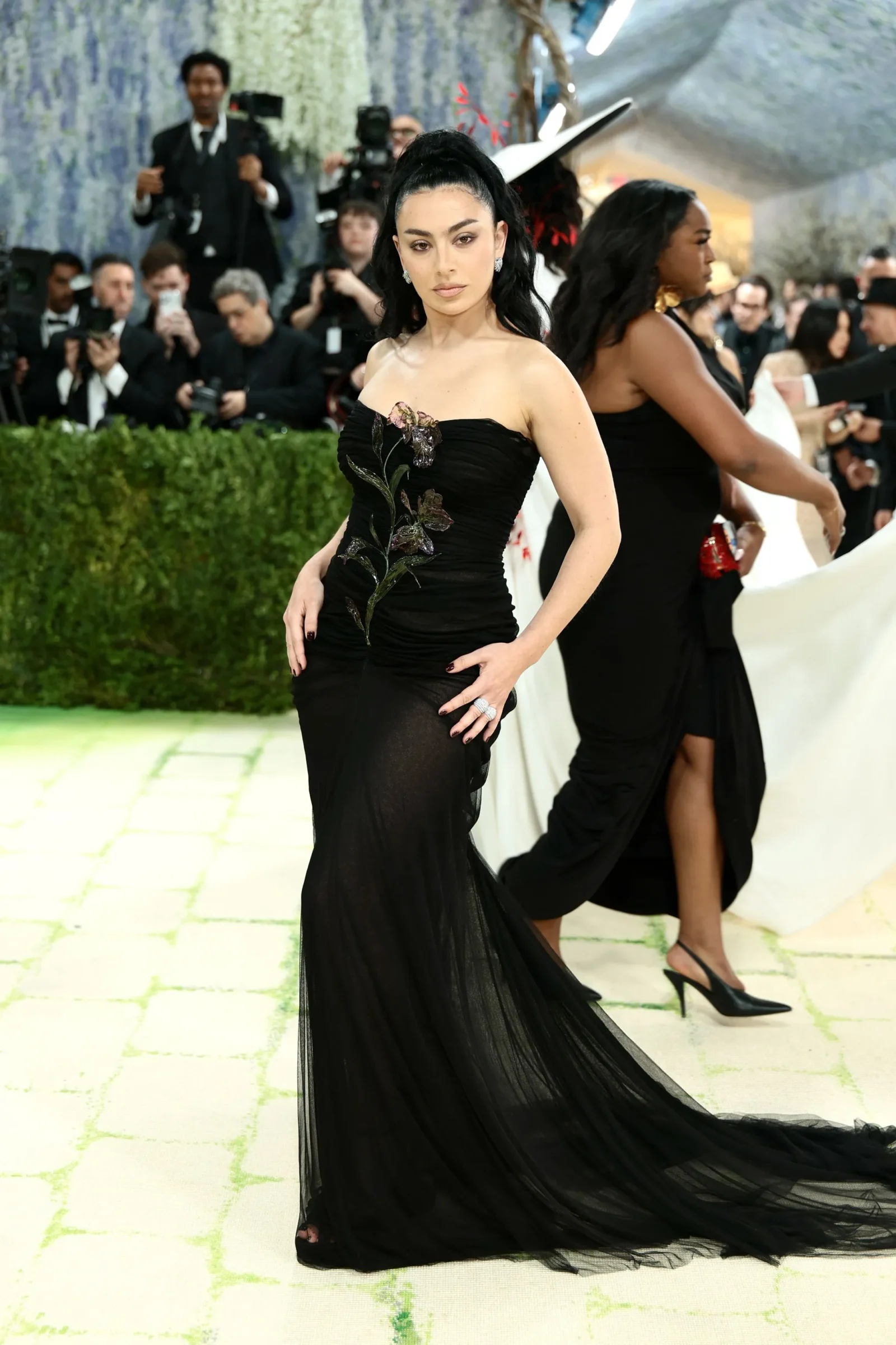 Woman on a red carpet wearing a black strapless gown with embroidered floral design, posing for photographers in the background.