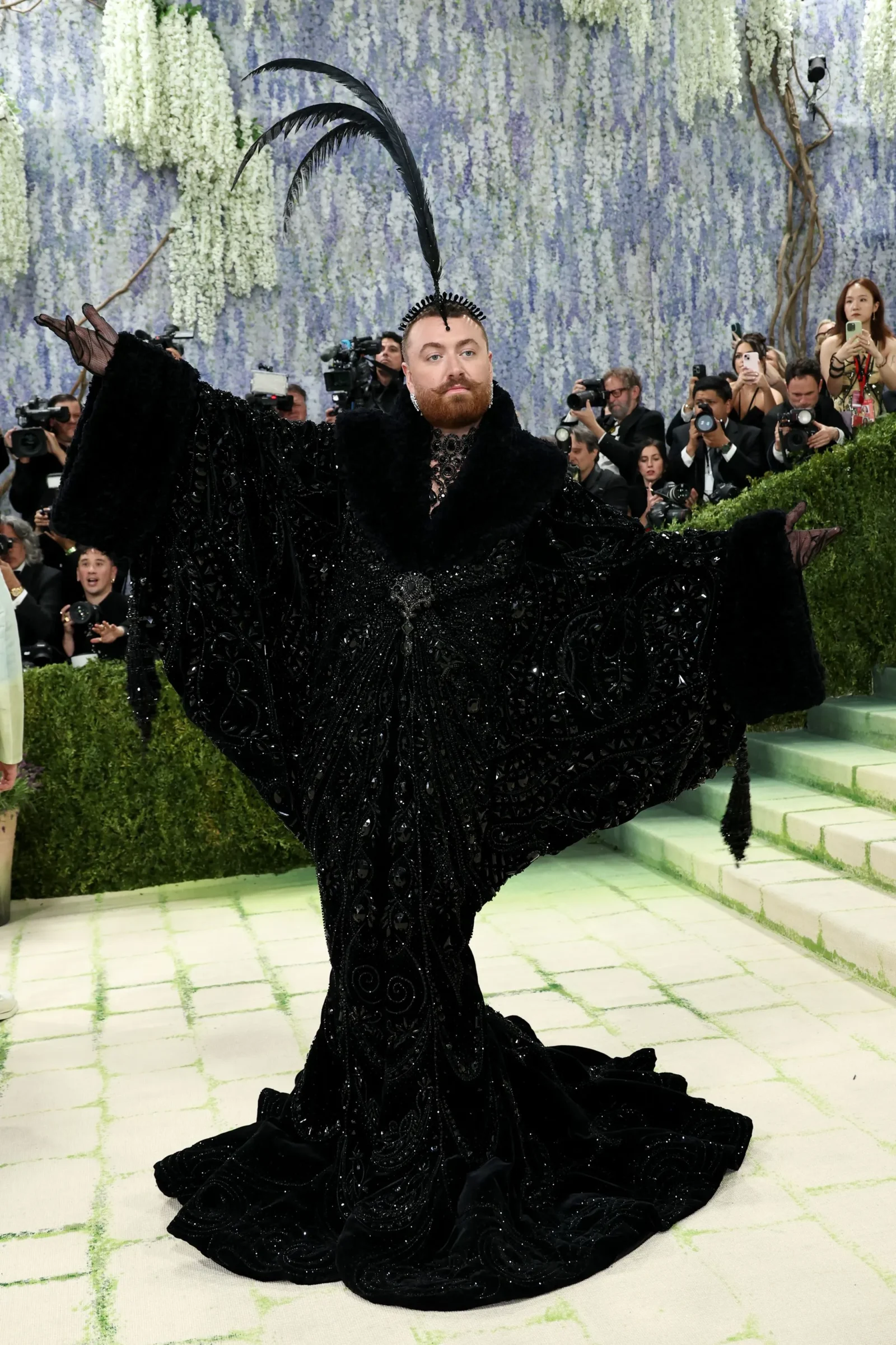 Person in a dramatic black gown with a tall feather headpiece posing for photographers at a gala.
