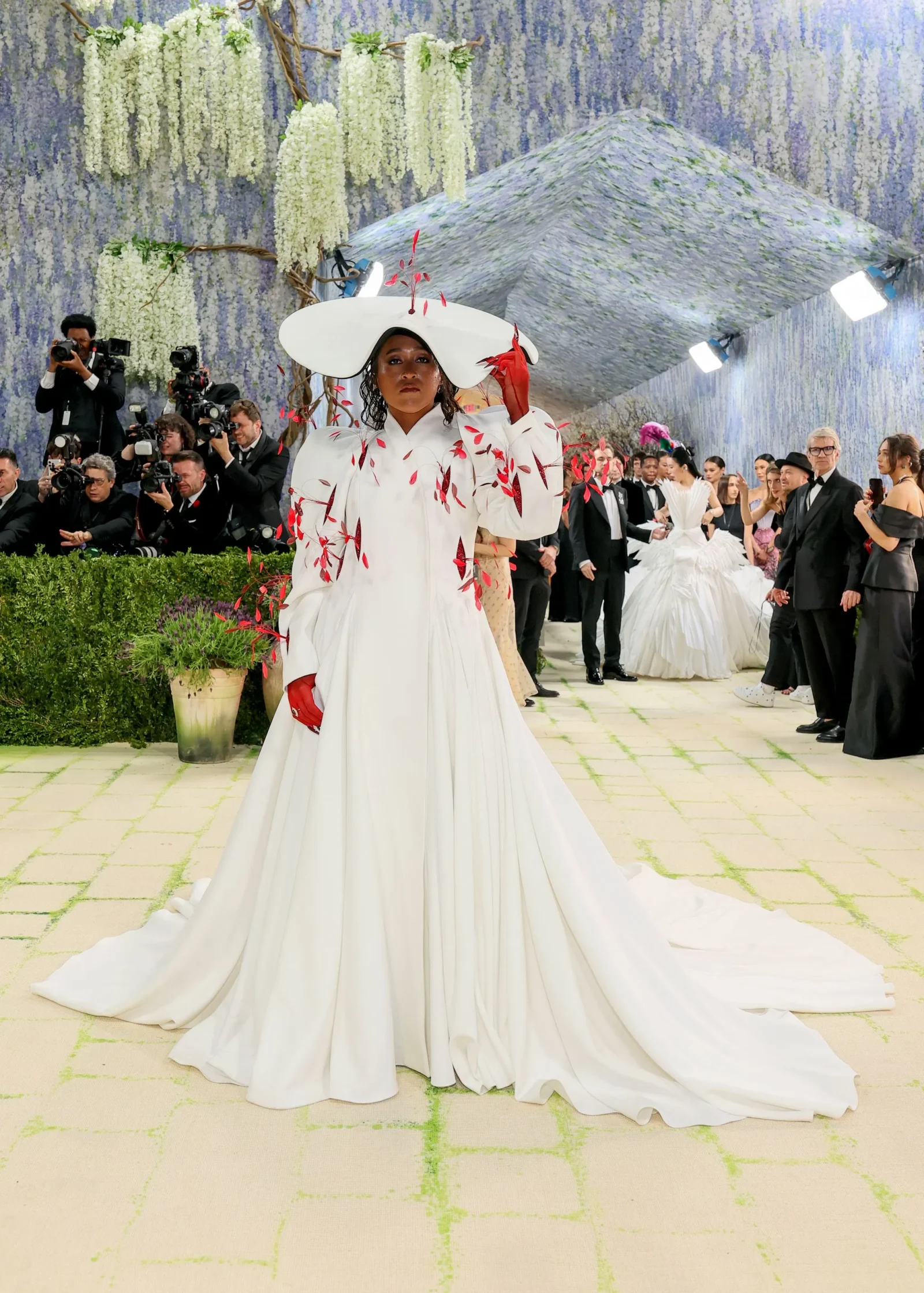 Model in a dramatic white couture gown with red leaf embroidery and a large white hat, posing for photographers on a floral backdrop.