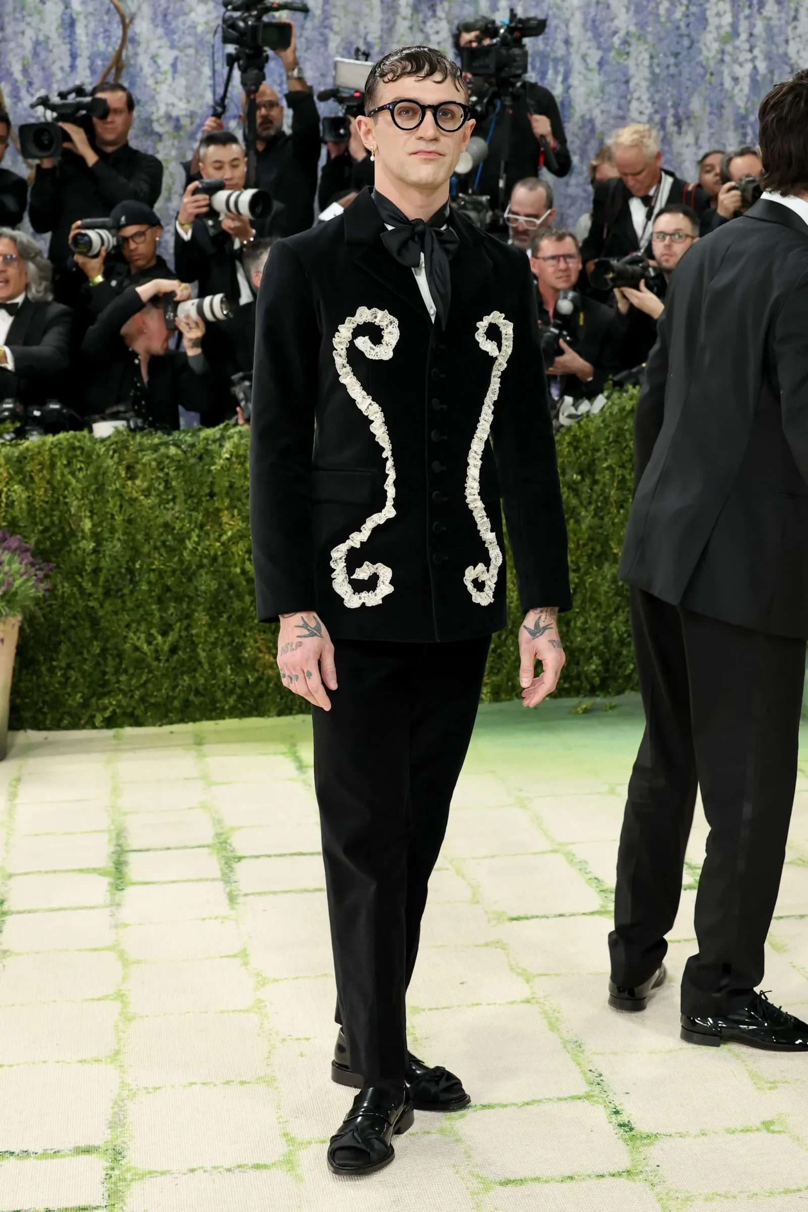 Man in a black velvet suit with white embroidery and large round glasses, standing on a patterned floor as photographers shoot in the background to his left and right.