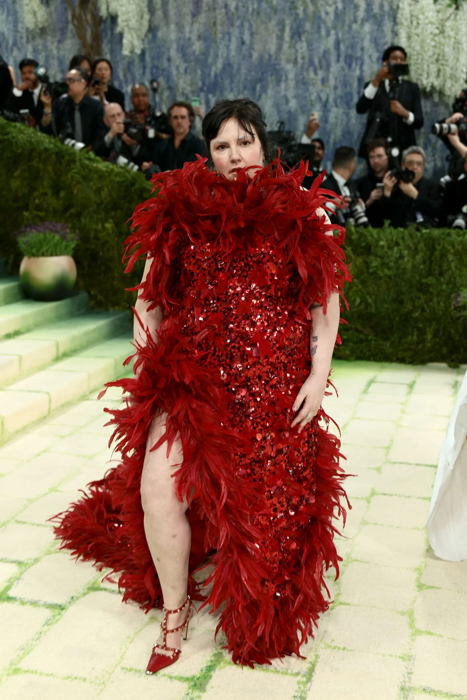 Woman in a red feathered, sequined gown posing on a light green carpet with photographers in the background