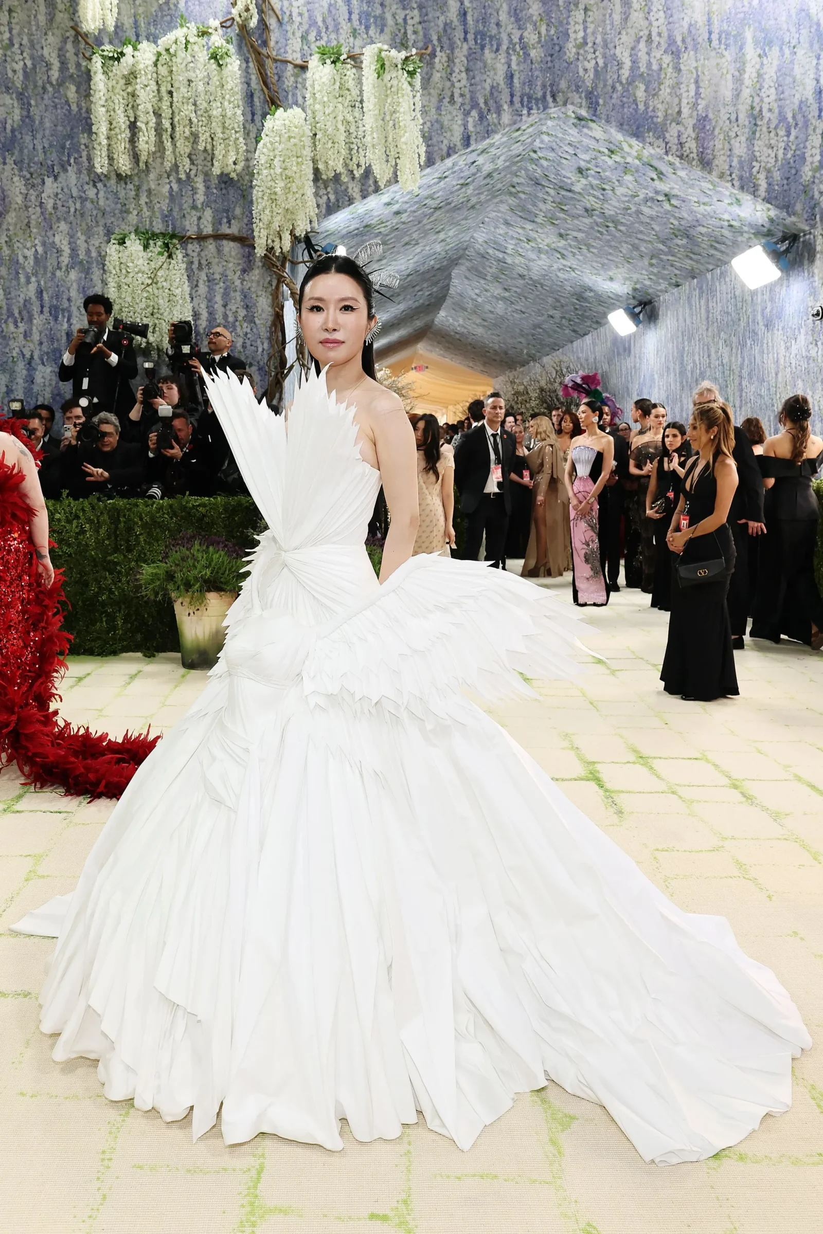 Elegantly dressed woman in a voluminous white gown on a red carpet, surrounded by photographers and guests under floral decor.