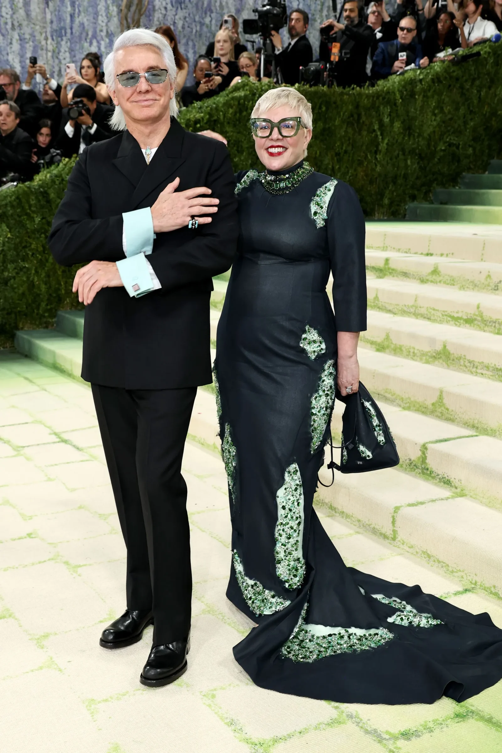 Two stylish celebrities pose on a light-green carpeted step; the man wears a black suit with pale blue cuffs and sunglasses, the woman in a dark gown with emerald beading and a long train, photographers in the background.