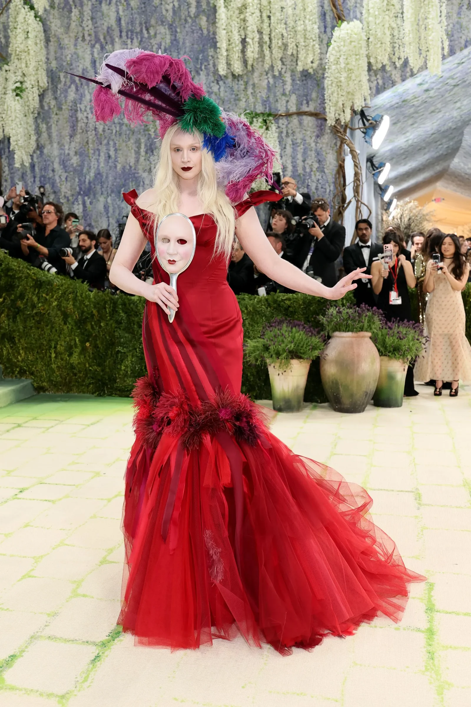 Woman in a dramatic red gown with a large feather headdress, holding a handheld mirror with a pale mask, on a green carpet with photographers in background.
