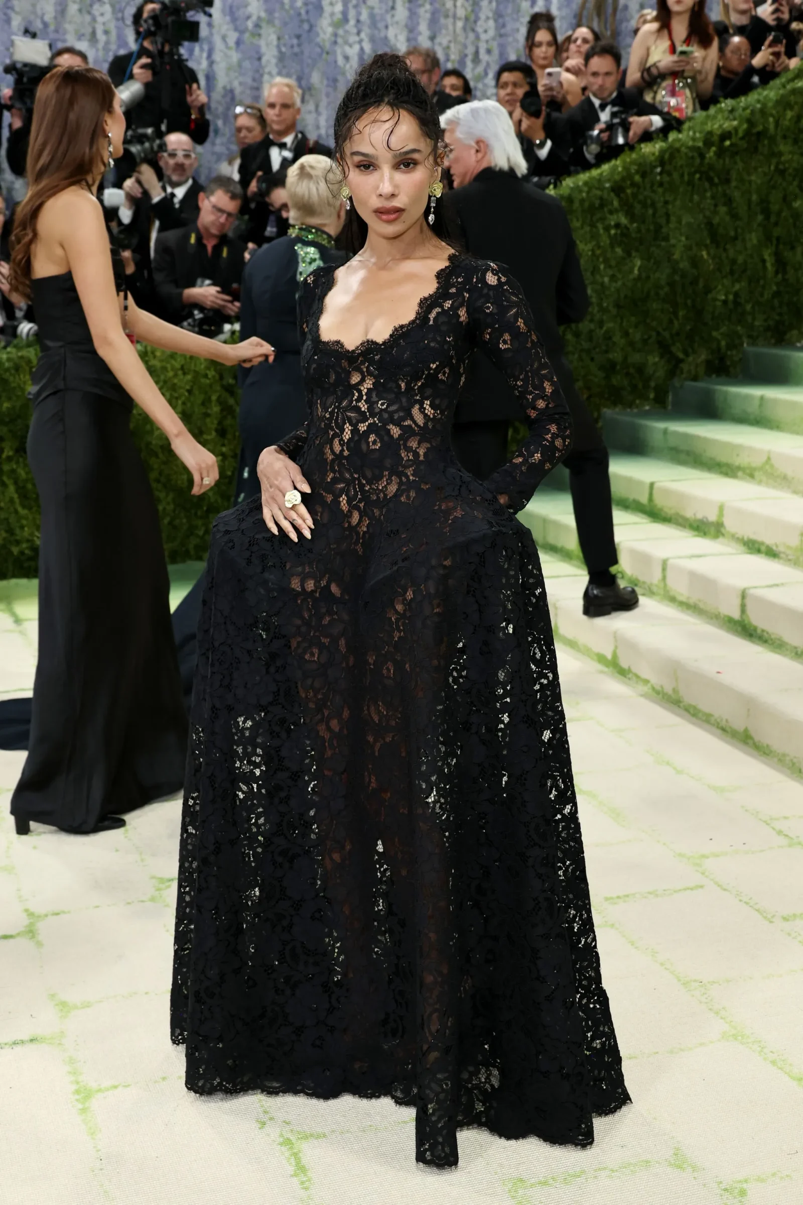 Woman in a black lace gown posing on a step red carpet, photographers in background.