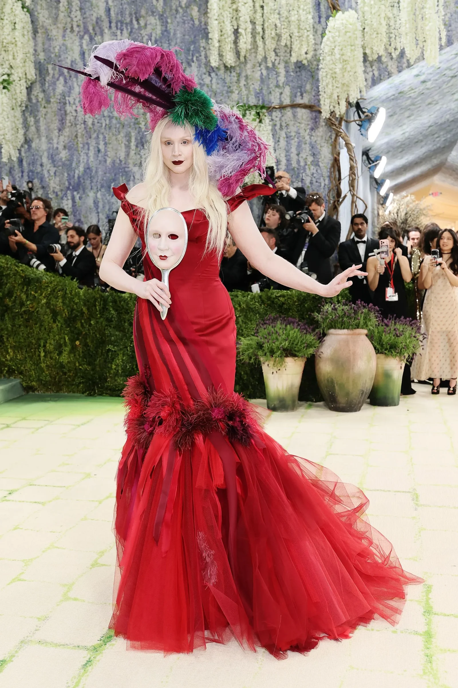 Model in a bright red gown with a large multi-colored feather headdress, holding a white mask, amid photographers at a glamorous event