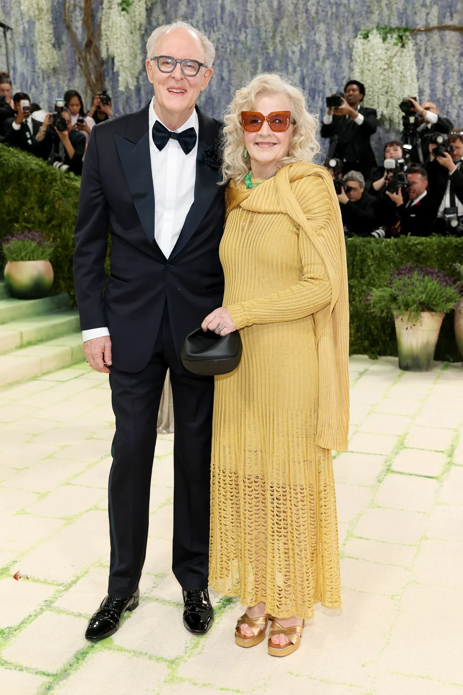 Elderly man in a black tuxedo with bow tie stands beside an elderly woman in a gold pleated dress and orange sunglasses in a formal event setting? photographers in the background.
