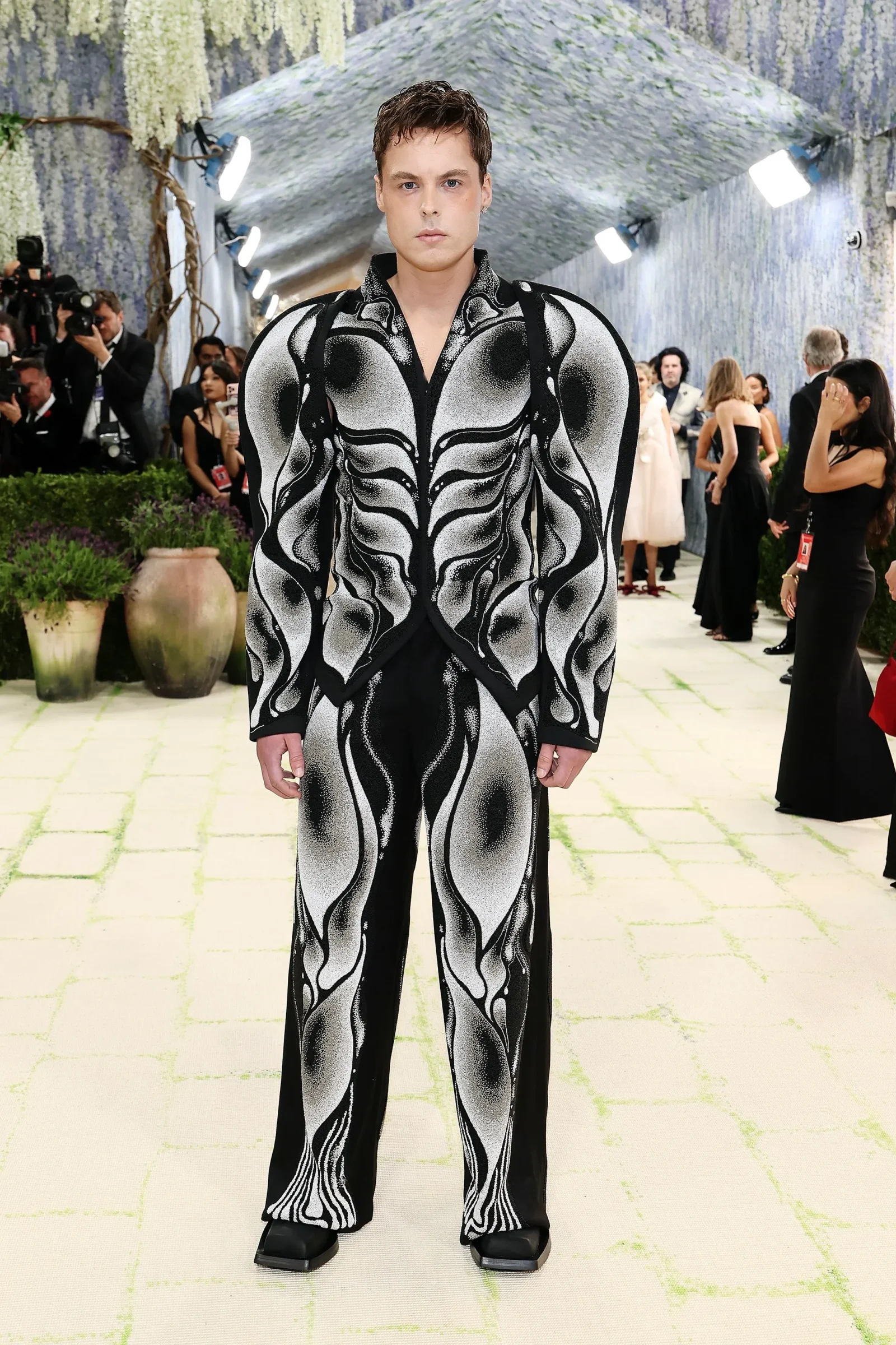 Male model wearing a bold black-and-white skeleton-pattern suit poses at a fashion event with photographers in the background.