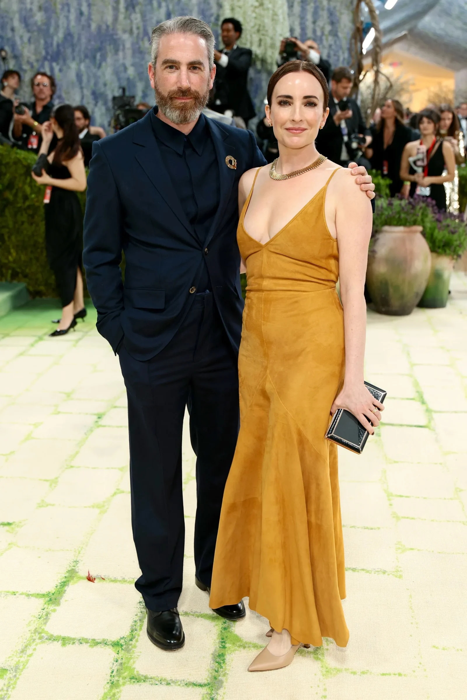 Smiling couple posing at a formal event: man in a navy suit and beard, woman in a mustard satin dress, holding a black clutch.