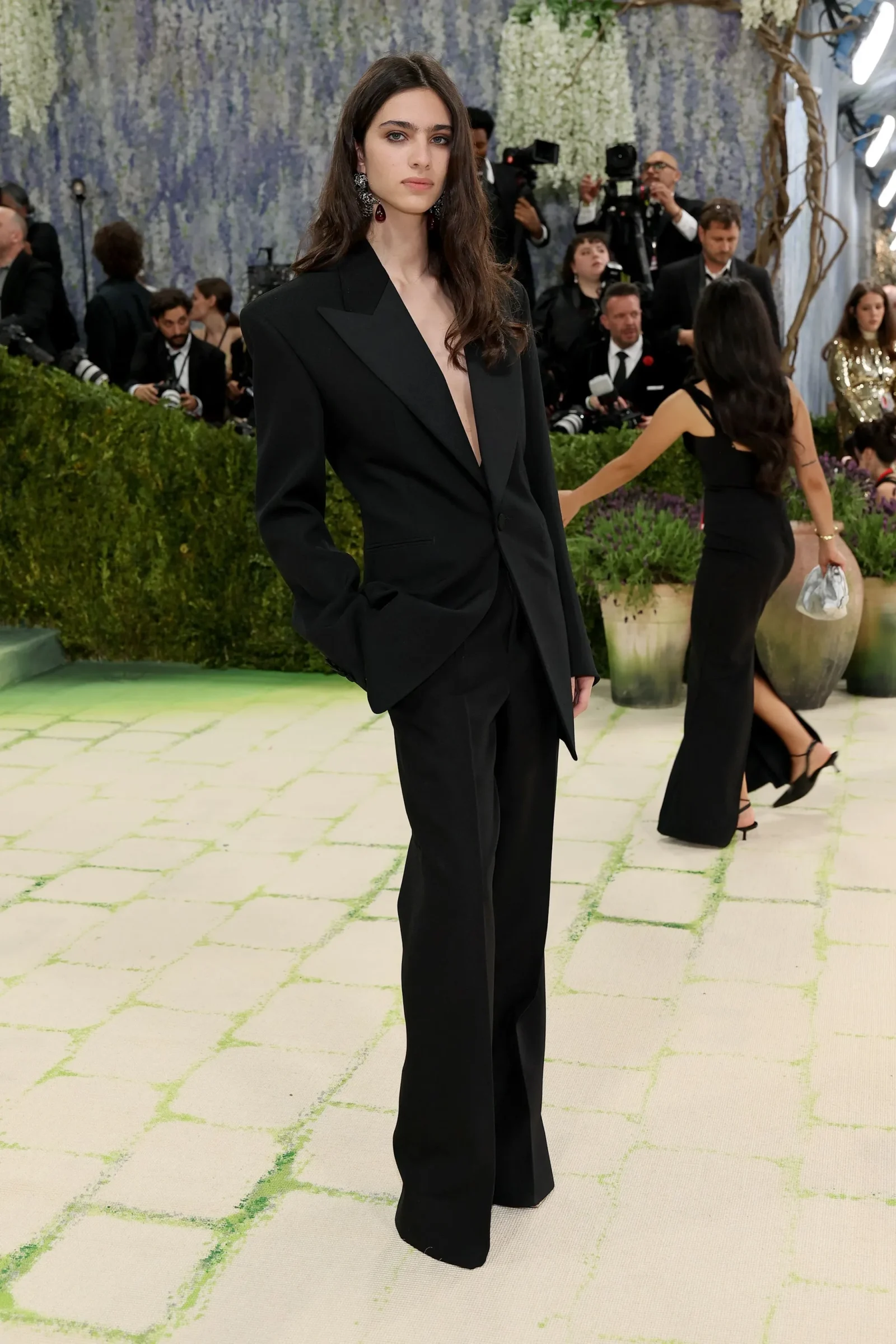 Young woman in a black tailored pantsuit posing on a pale green grid floor at a formal event, with photographers in the background.