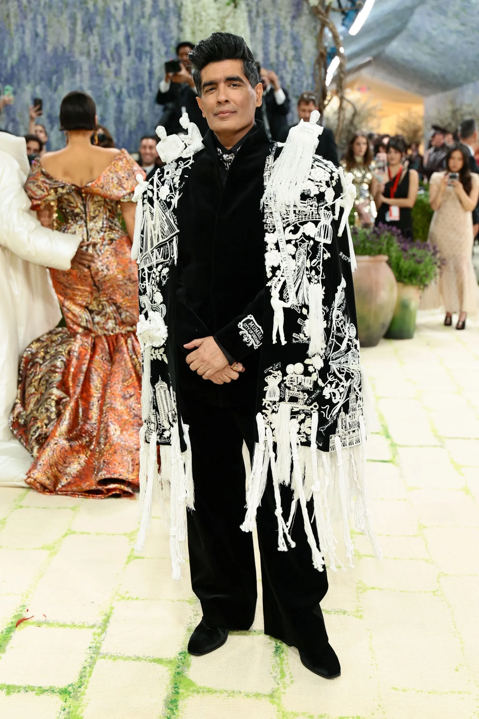 Man in a black suit with a detailed white embroidered cape posing at a formal event, with attendees in gowns in the background.