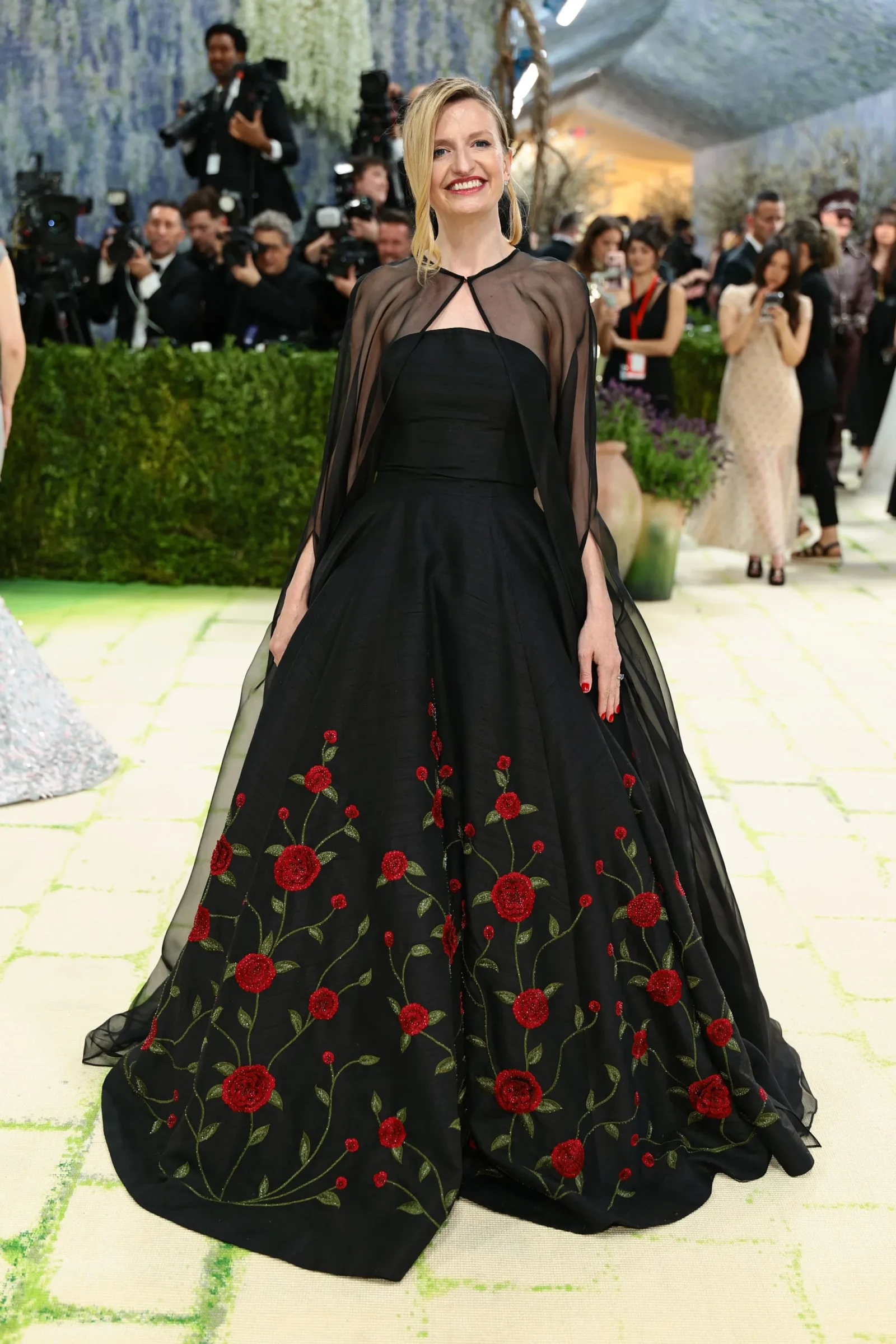 Woman in a black, floor-length gown with sheer cape and red floral embroidery posing on a light-green carpet as photographers line the background.