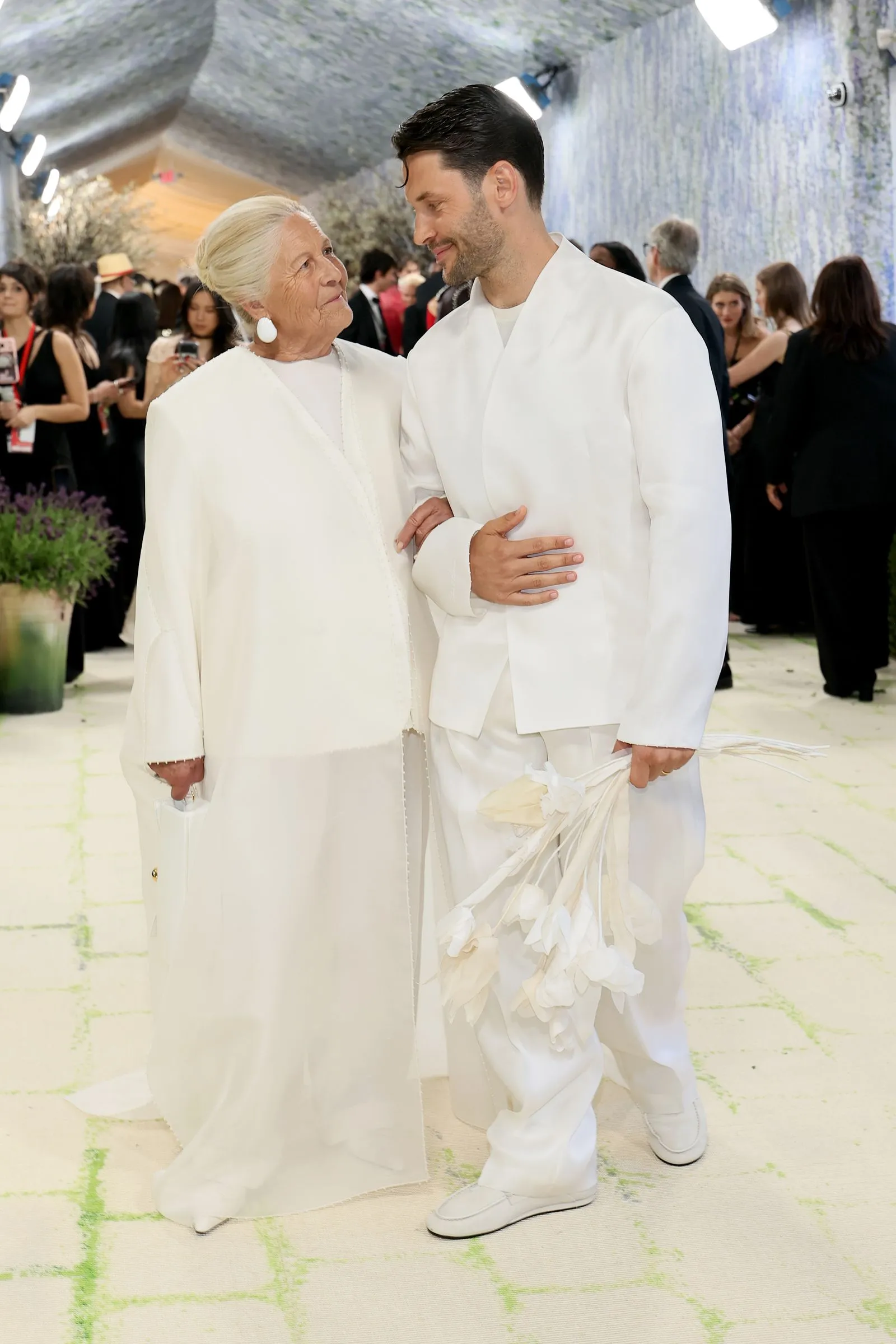 Two people in all-white outfits, an elderly woman and a younger man, walk arm in arm at a formal event with a crowd in the background.