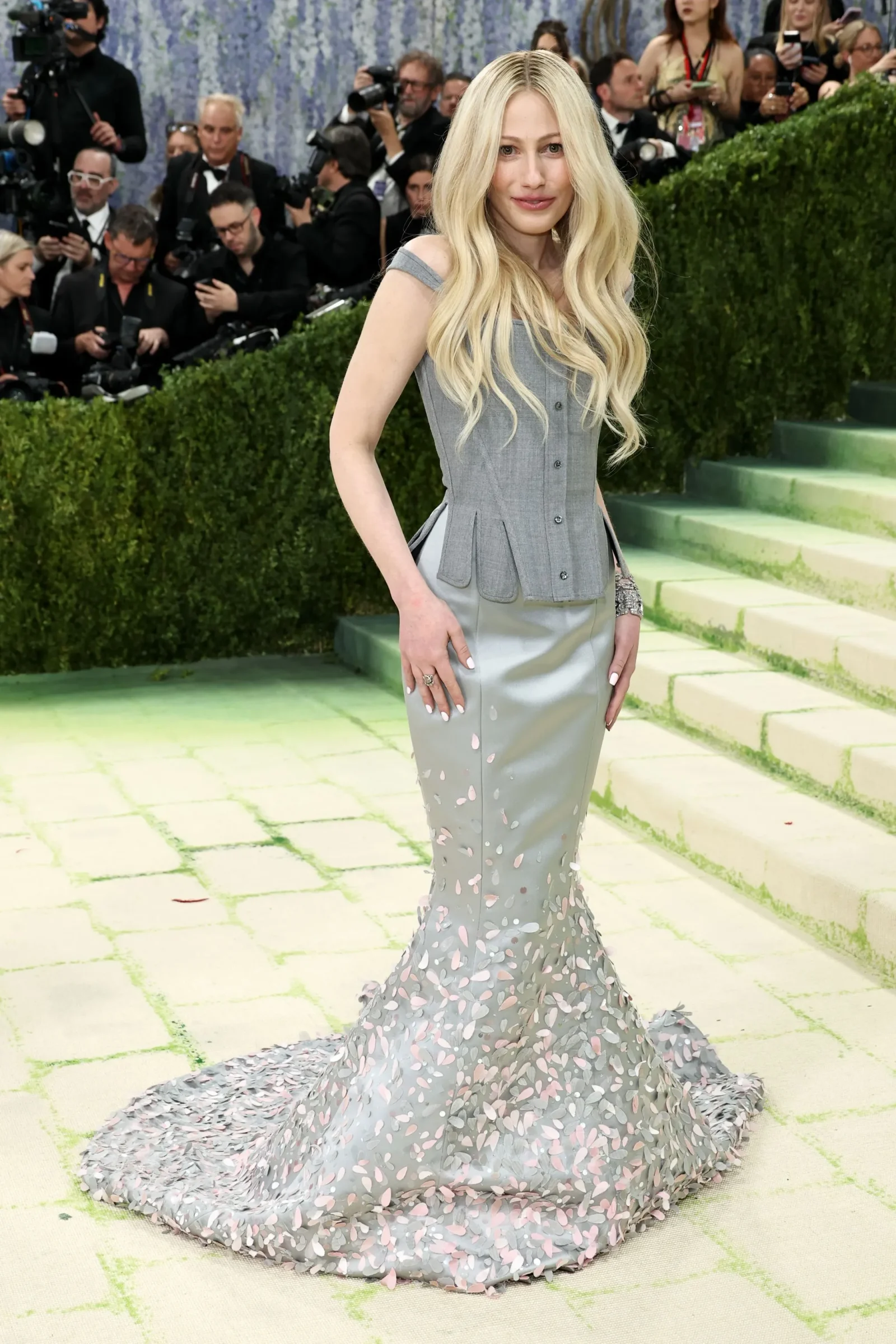 Blonde woman in a gray gown with petal appliqués posing on a green-toned carpet while photographers crowd behind her