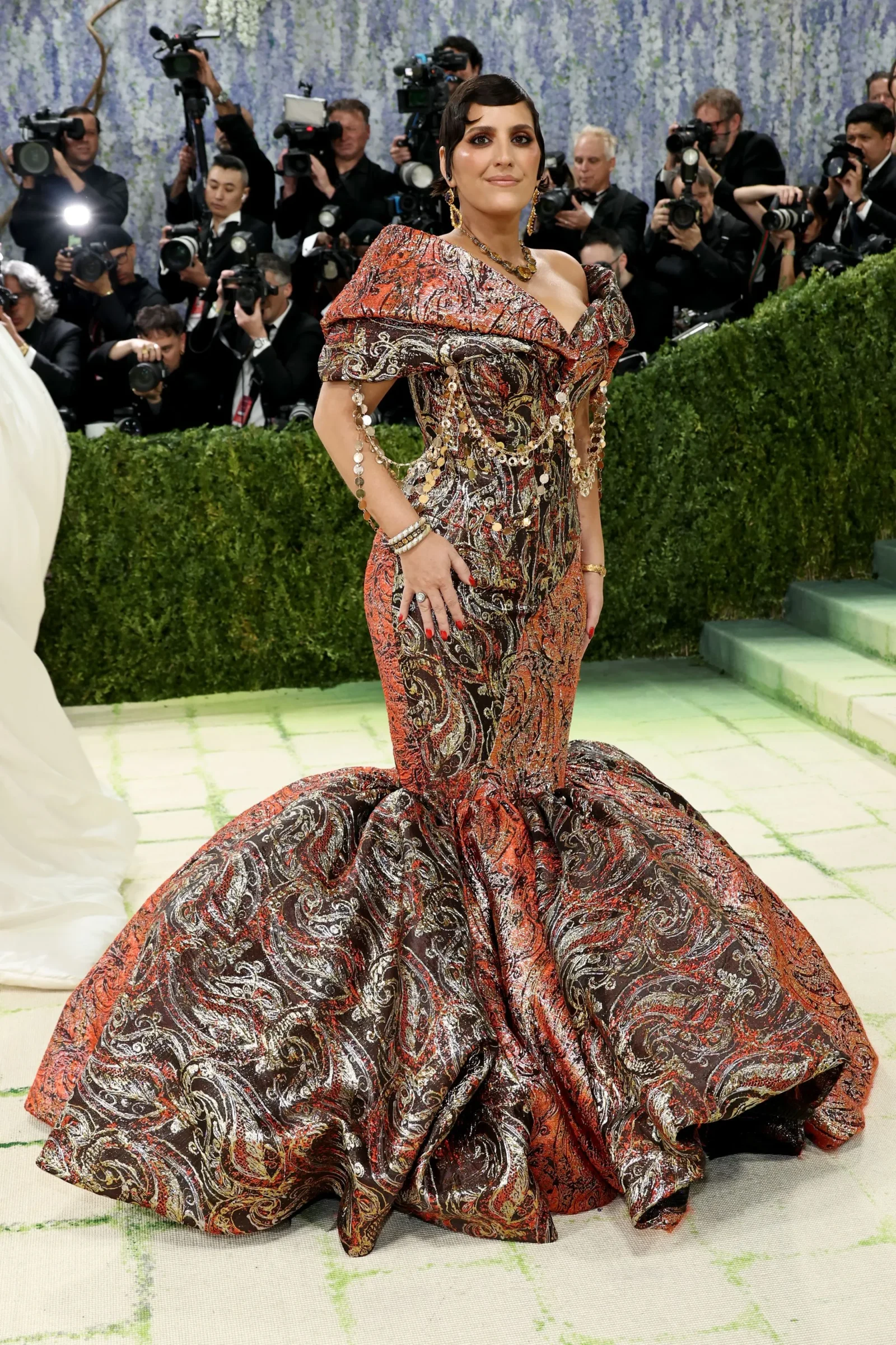 Celebrity on a red carpet wearing an ornate red-and-black patterned gown with jewelry, posing for photographers in the background.