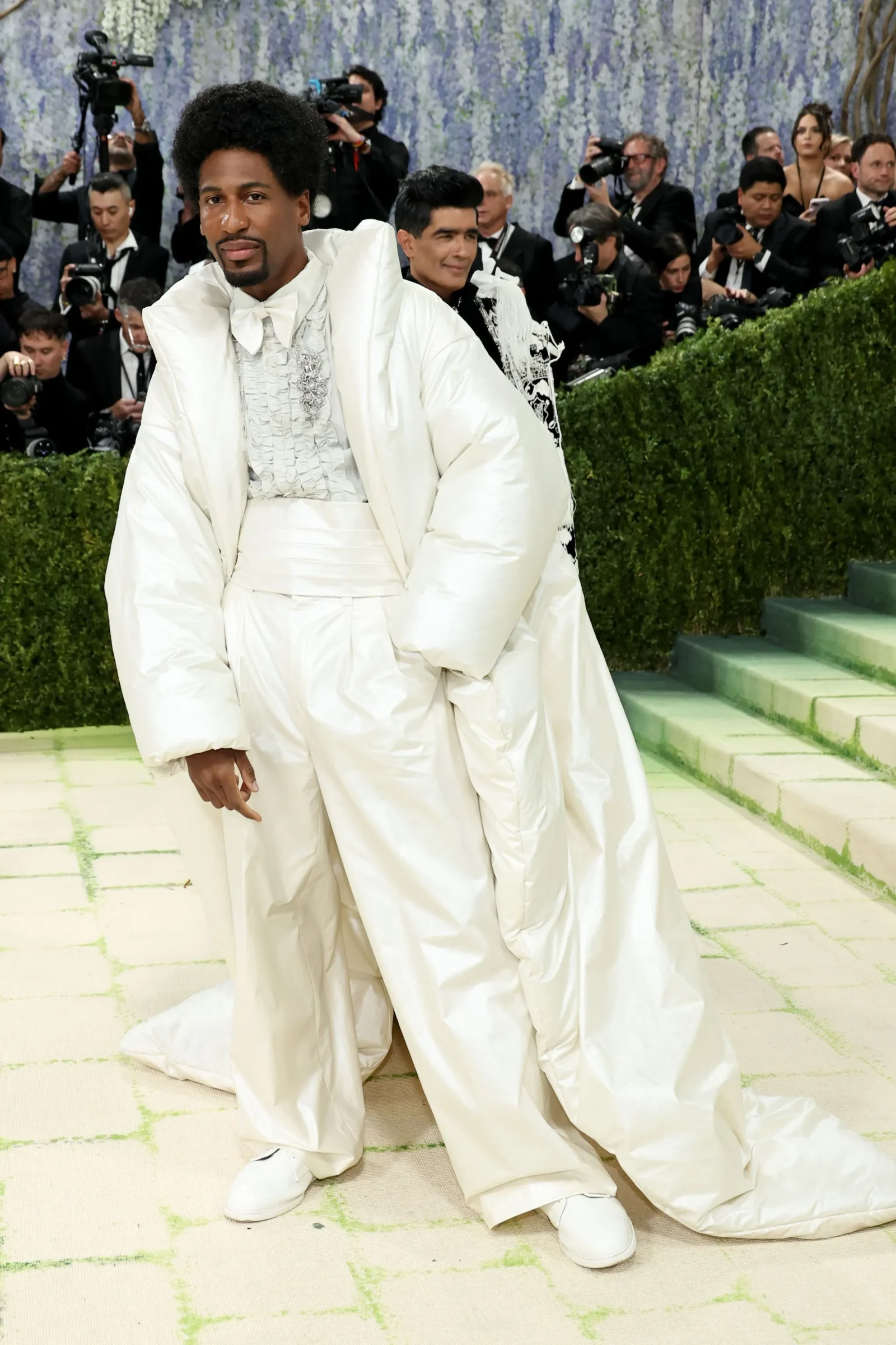Man in an oversized white suit posing on a pale carpet as photographers line the background.