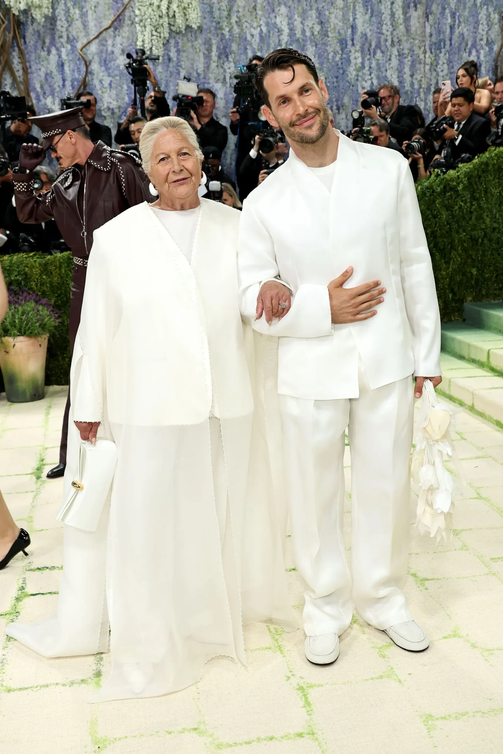 Older woman and younger man pose together in white outfits at a high-profile event, surrounded by photographers on a floral backdrop.