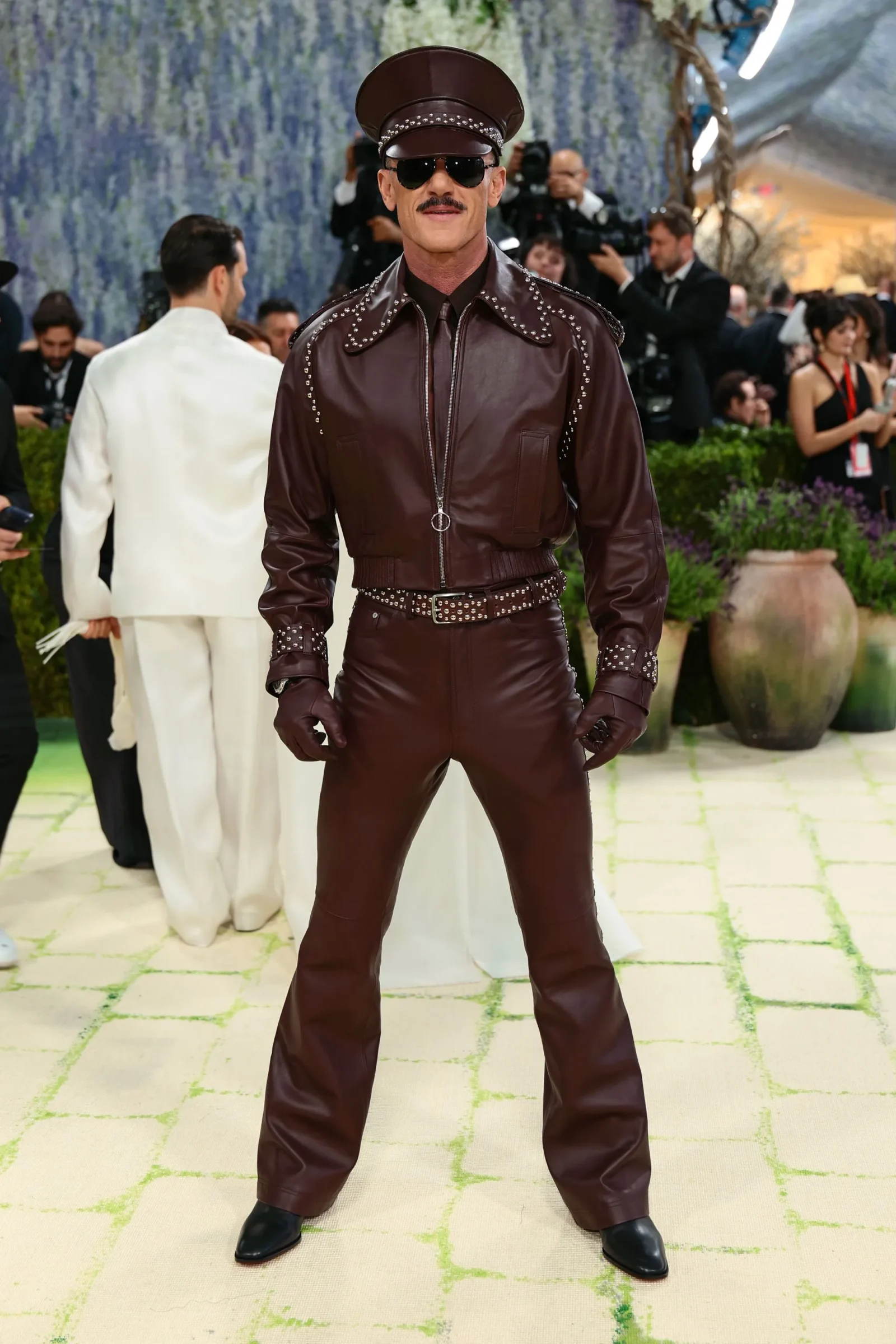 Man in brown studded leather outfit and matching cap posing on a light patterned carpet with photographers in the background.