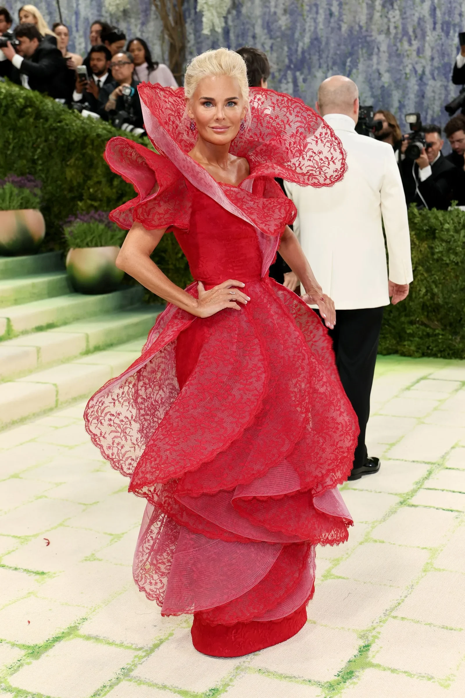Woman in a dramatic red lace gown posing on a light-green carpet at a crowded event with photographers in the background, front and center.