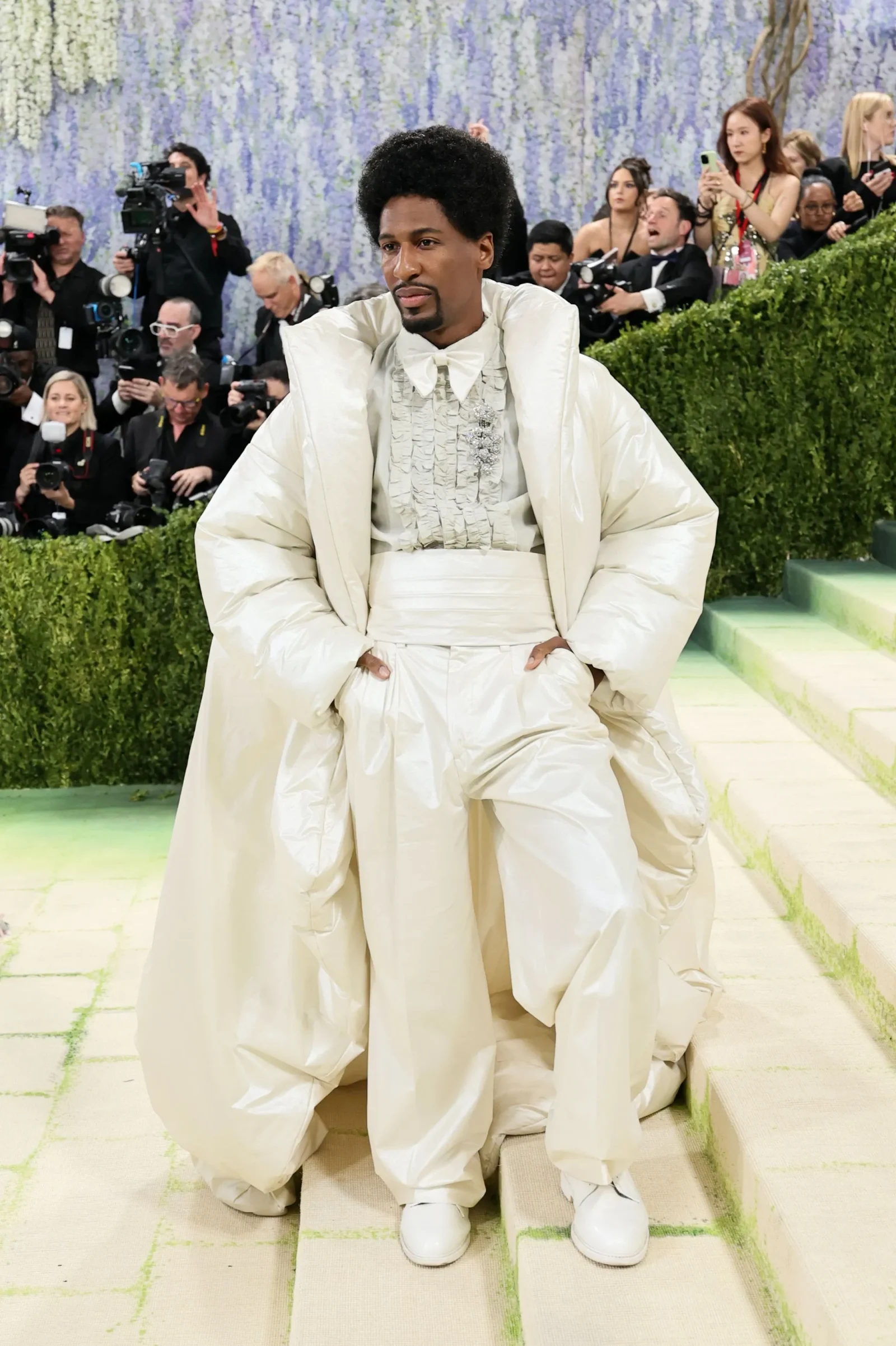 Man in a voluminous white couture outfit posing on steps as photographers film him in the background.