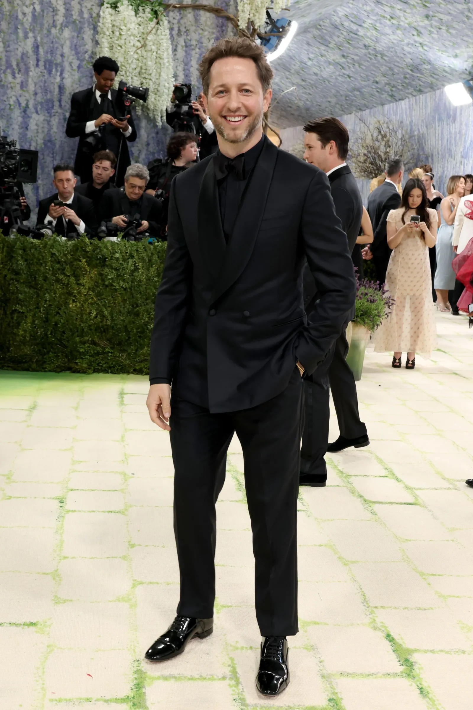 Smiling man in a black tuxedo posing on a light tiled floor with photographers and guests in the background.